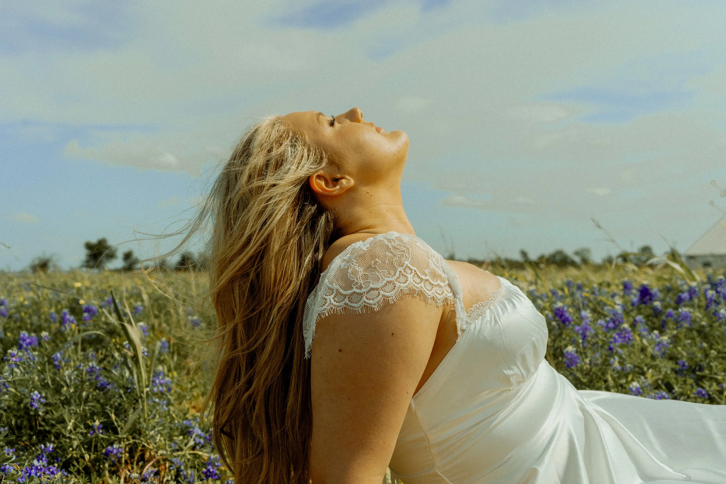 A woman with long blonde hair and fair skin wearing a white lace top, leaning back with her face tilted upward in a field of purple and yellow wildflowers under a partly cloudy sky.