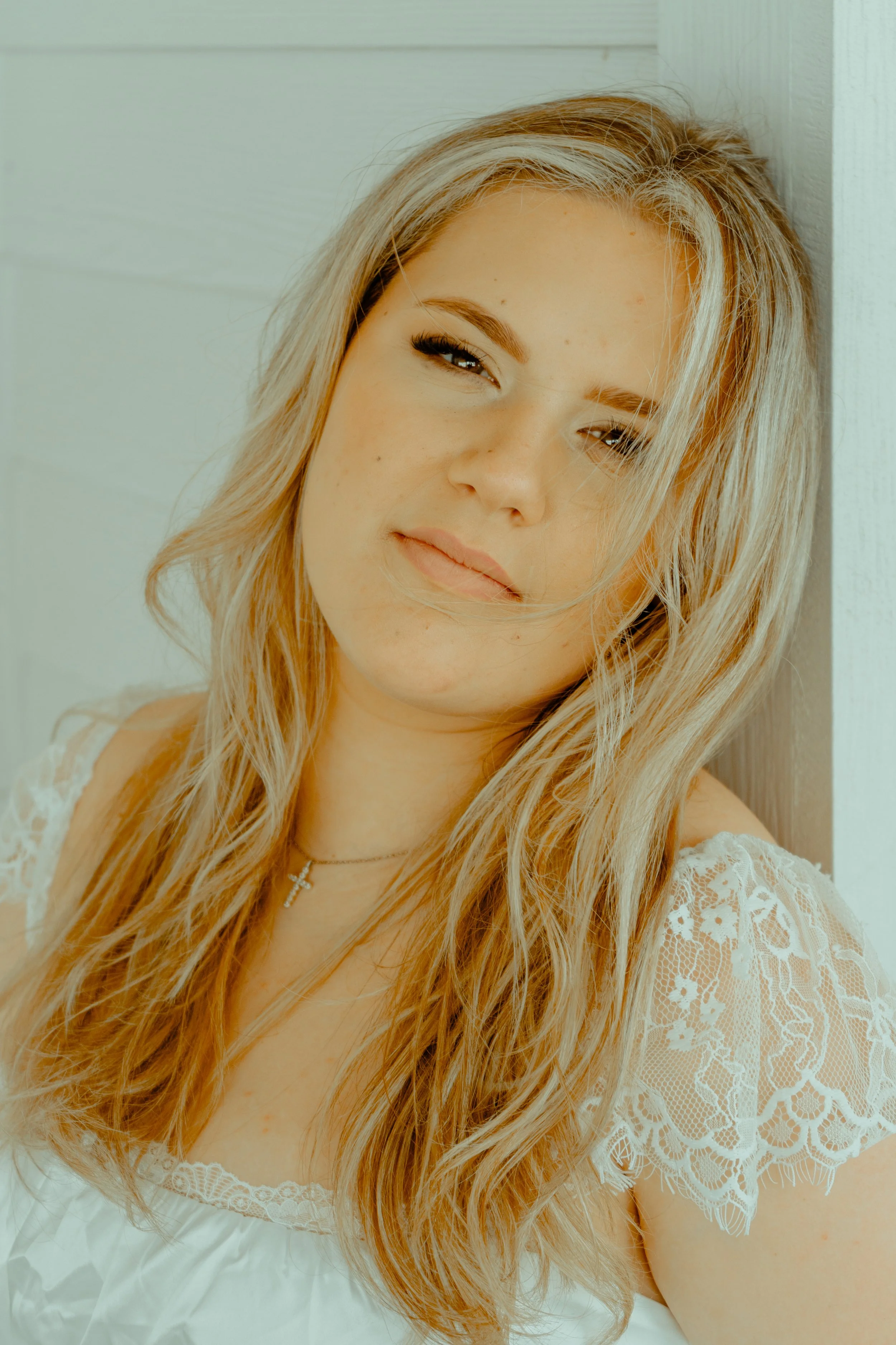 Close-up of a young woman with long wavy blonde hair, wearing a white lace dress and a cross necklace, leaning against a light-colored wooden wall.