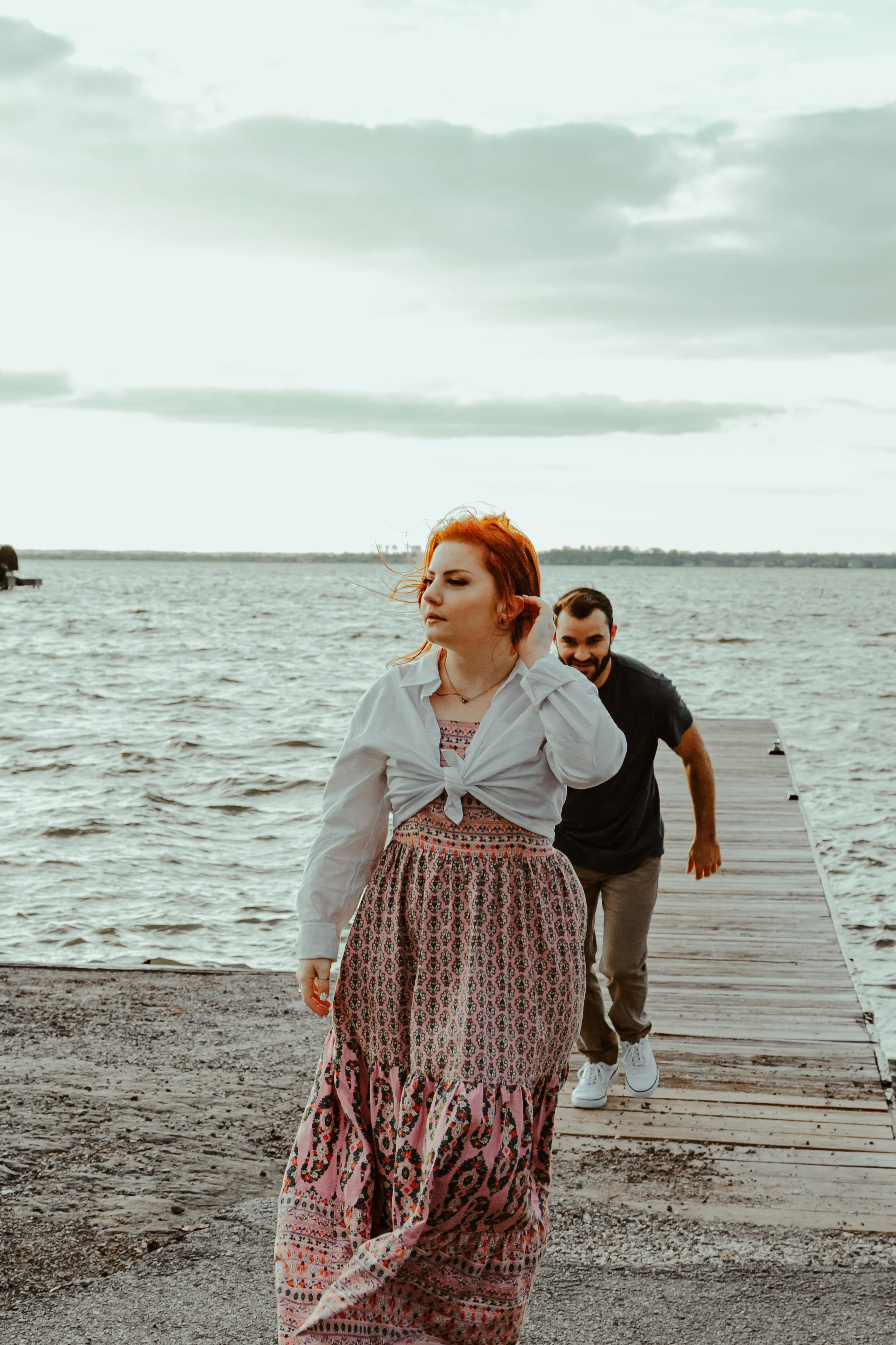 A woman in a white blouse and patterned long skirt walking on a wooden pier by a body of water, with a man behind her, all under a cloudy sky.