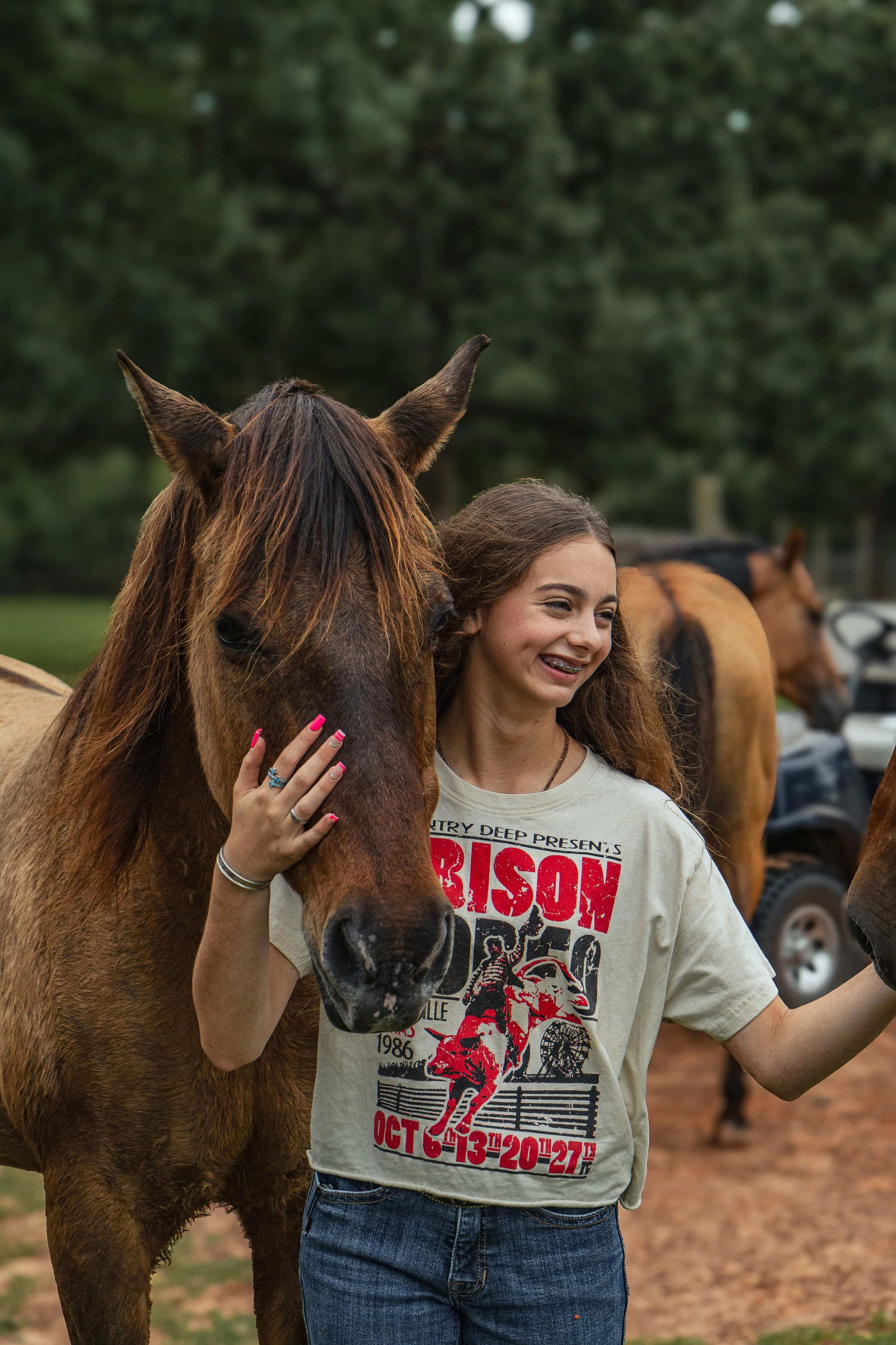 Young girl with wavy brown hair smiling and standing next to a brown horse, outdoors with trees in the background.