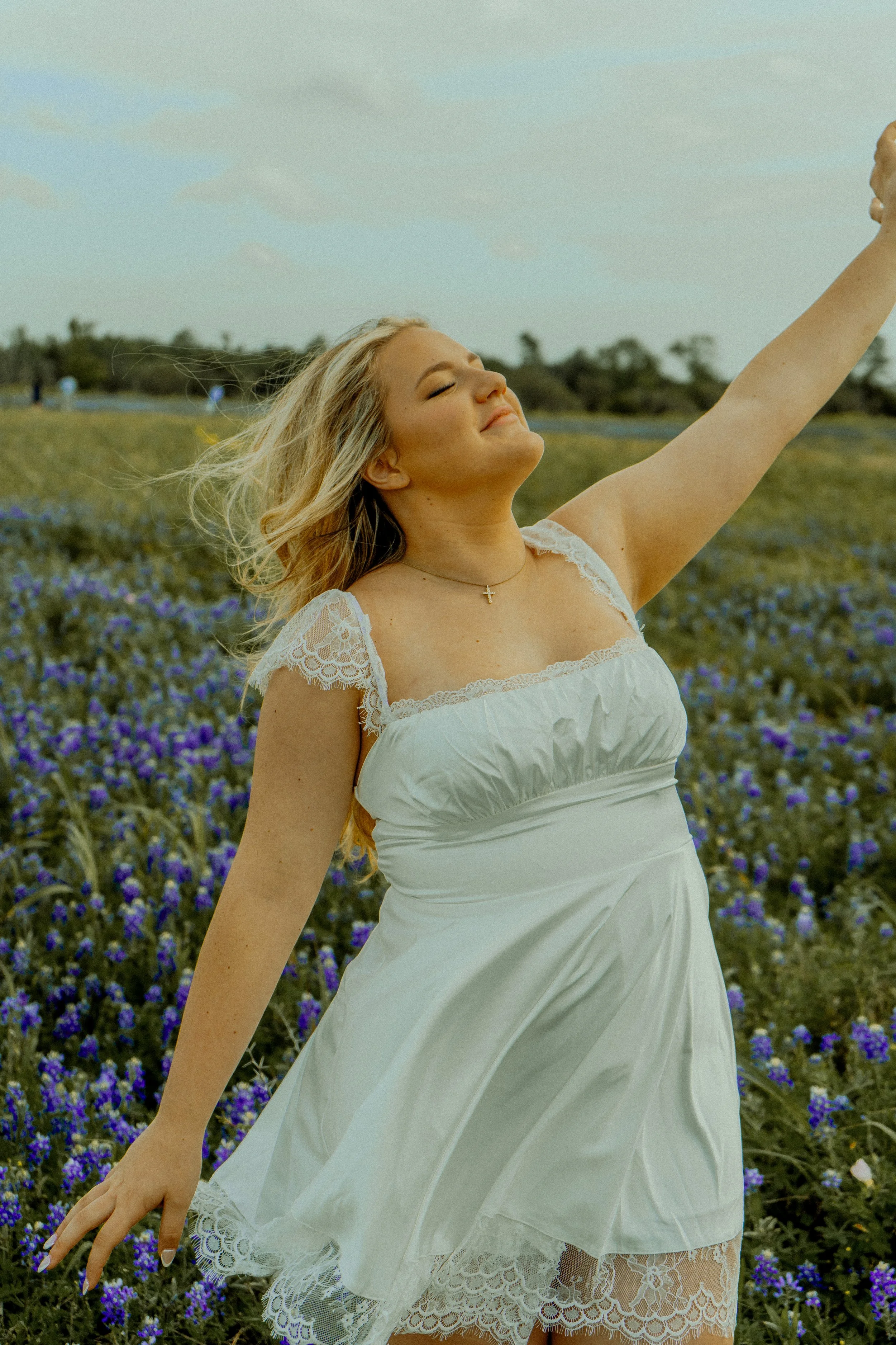 Young woman with blonde hair wearing a white dress with lace details, standing in a field of purple flowers with her eyes closed and arm raised, smiling.