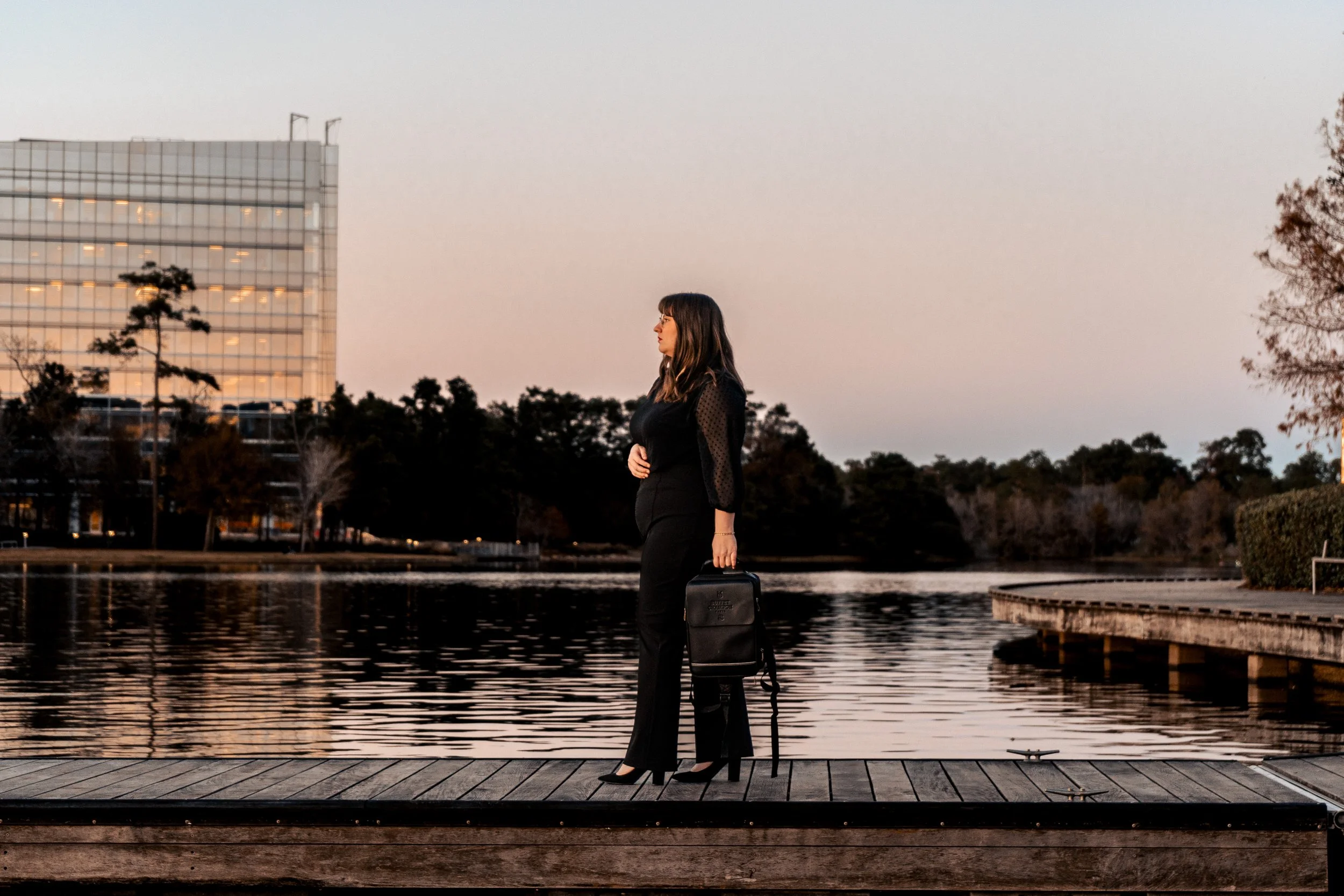 A woman in black clothing with a black handbag and high heels standing on a wooden dock by a lake during sunset, holding a suitcase, with trees and a modern glass building in the background.
