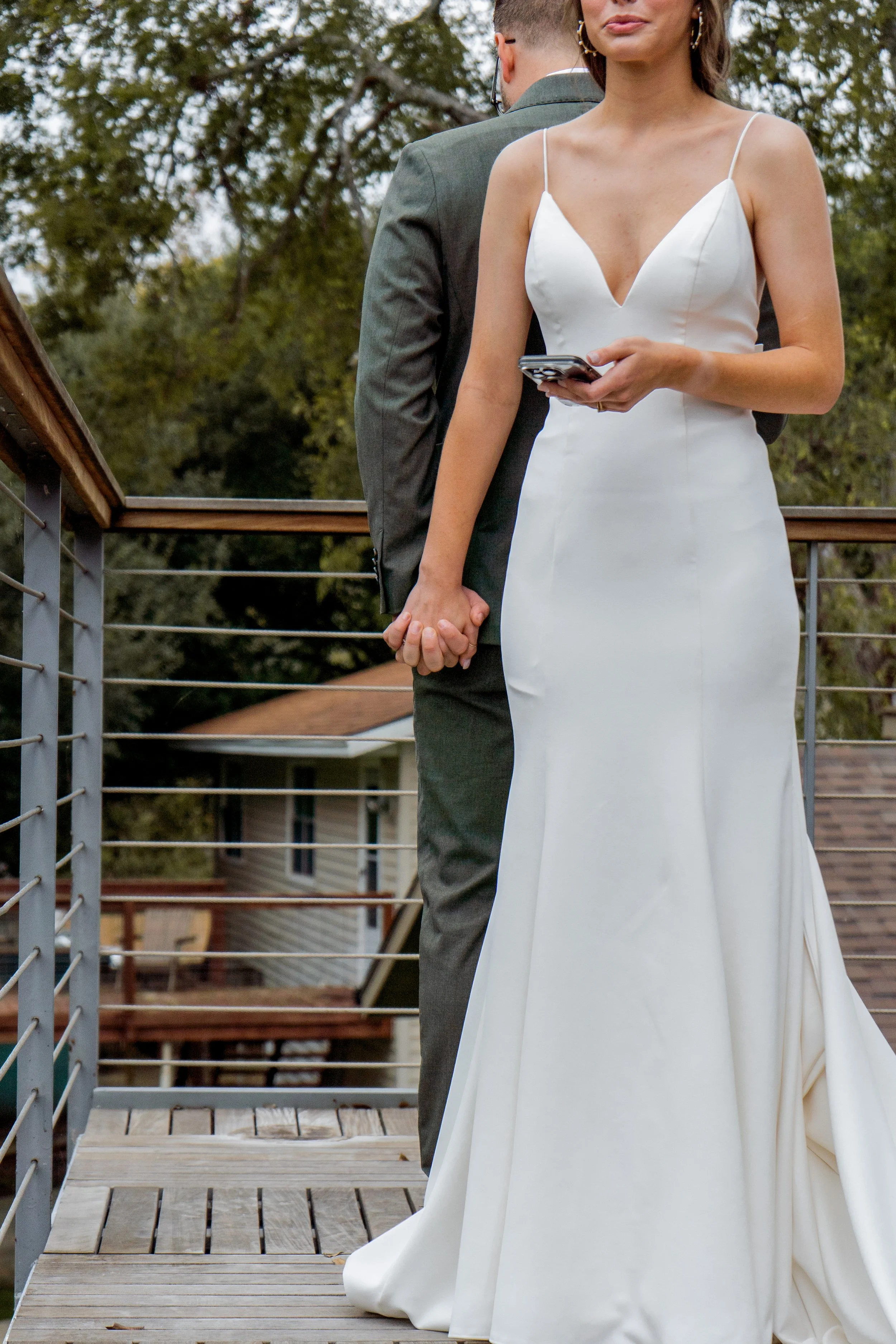 A bride in a white wedding dress holding hands with a groom in a gray suit during their private vows, standing on a wooden deck outdoors.