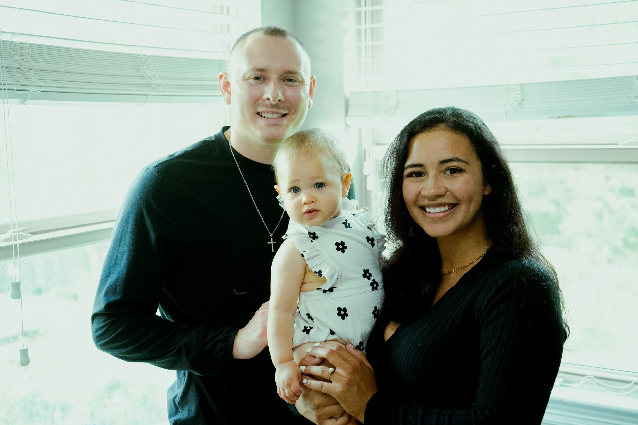 A smiling man in a black shirt holding a young girl with curly blonde hair and a white dress with black flowers, standing next to a smiling woman with long dark hair in an indoor setting with large windows.
