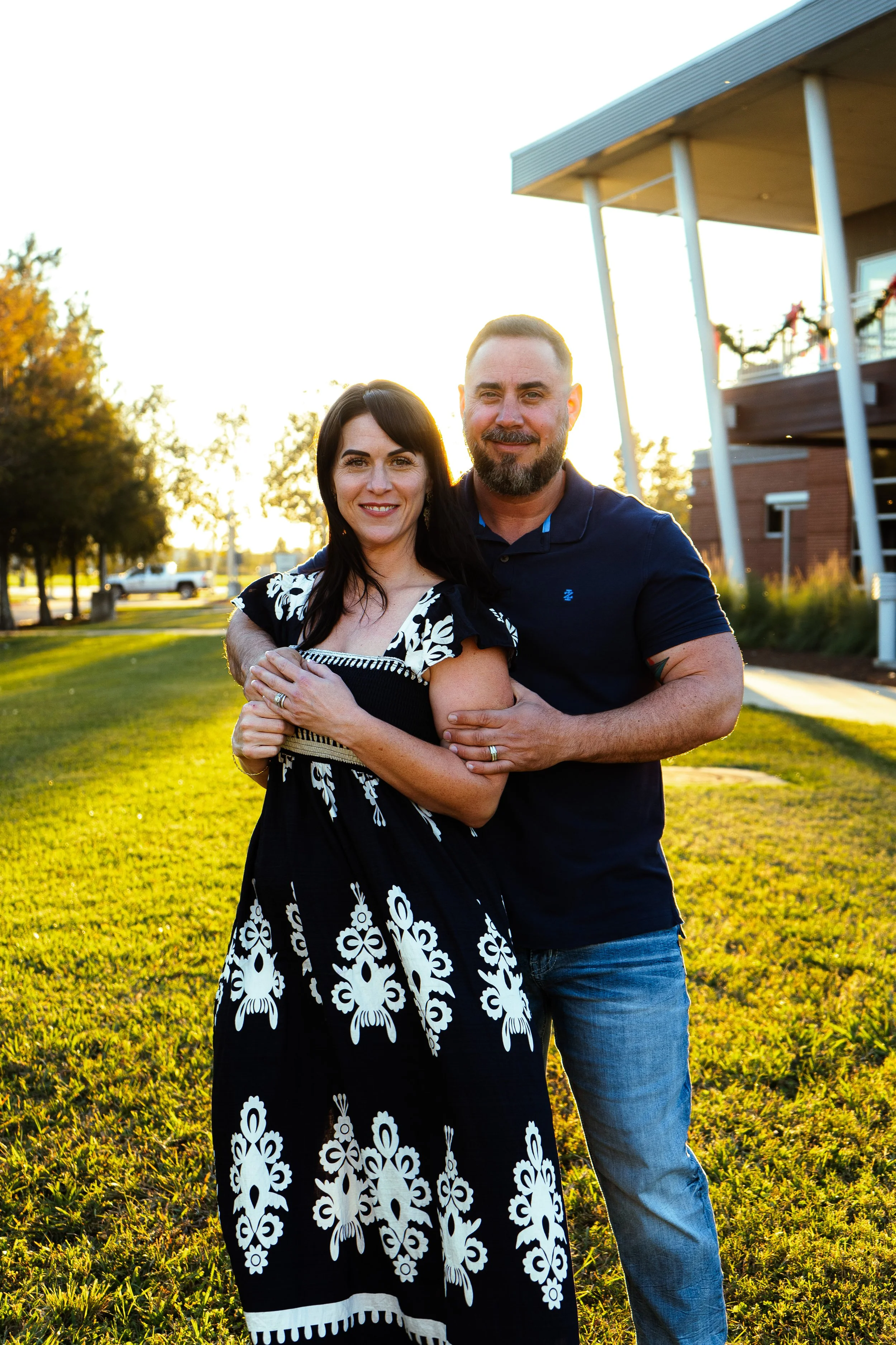 A smiling couple, a woman with dark hair in a patterned black and white dress, and a man with a beard in a navy shirt, standing outdoors during sunset on a grassy area with trees and a building in the background.