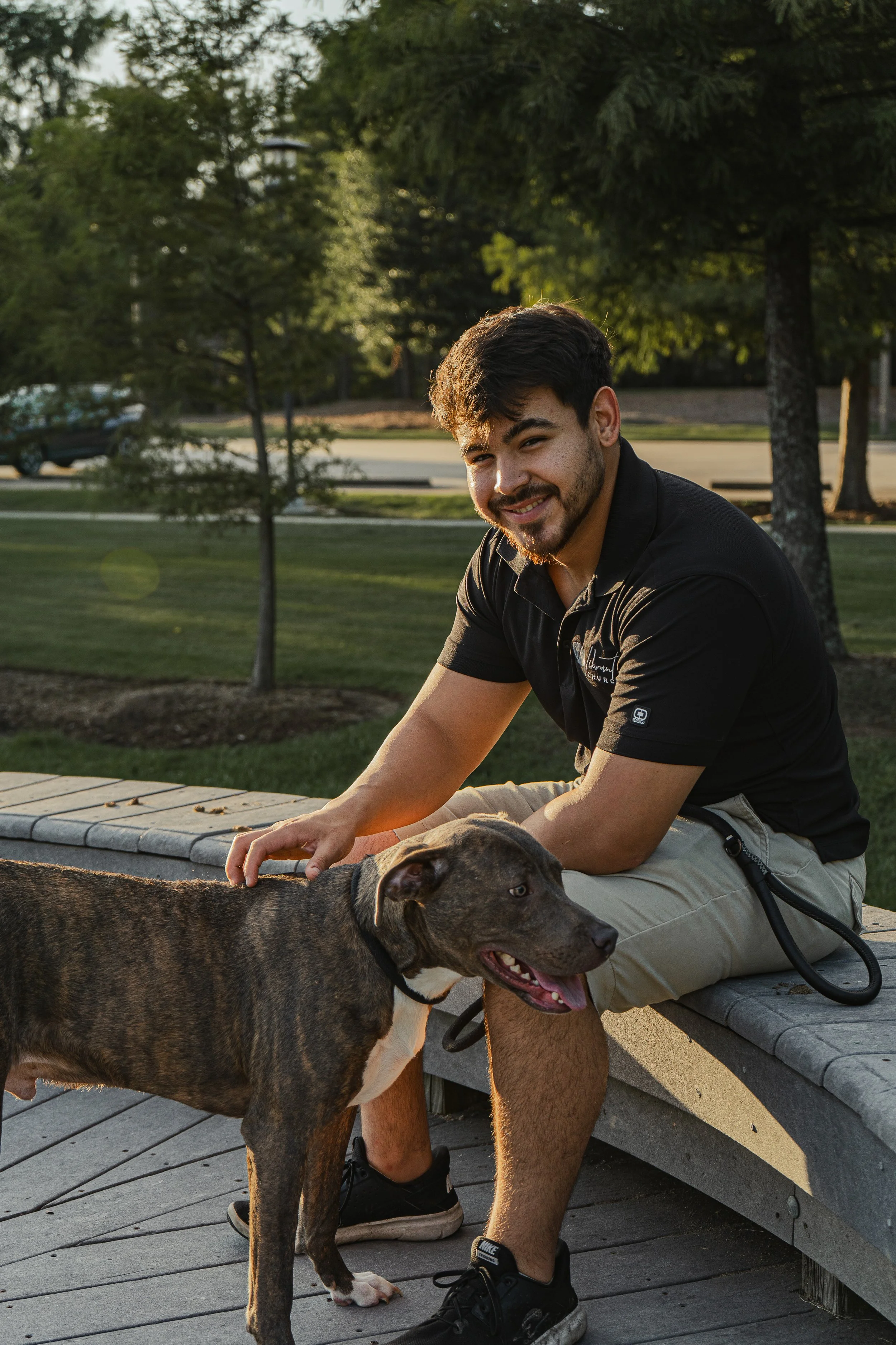 A young man with dark hair and a beard petting a large brindle dog outside in a park during the late afternoon or early evening. The man is sitting on a wooden bench with trees in the background.
