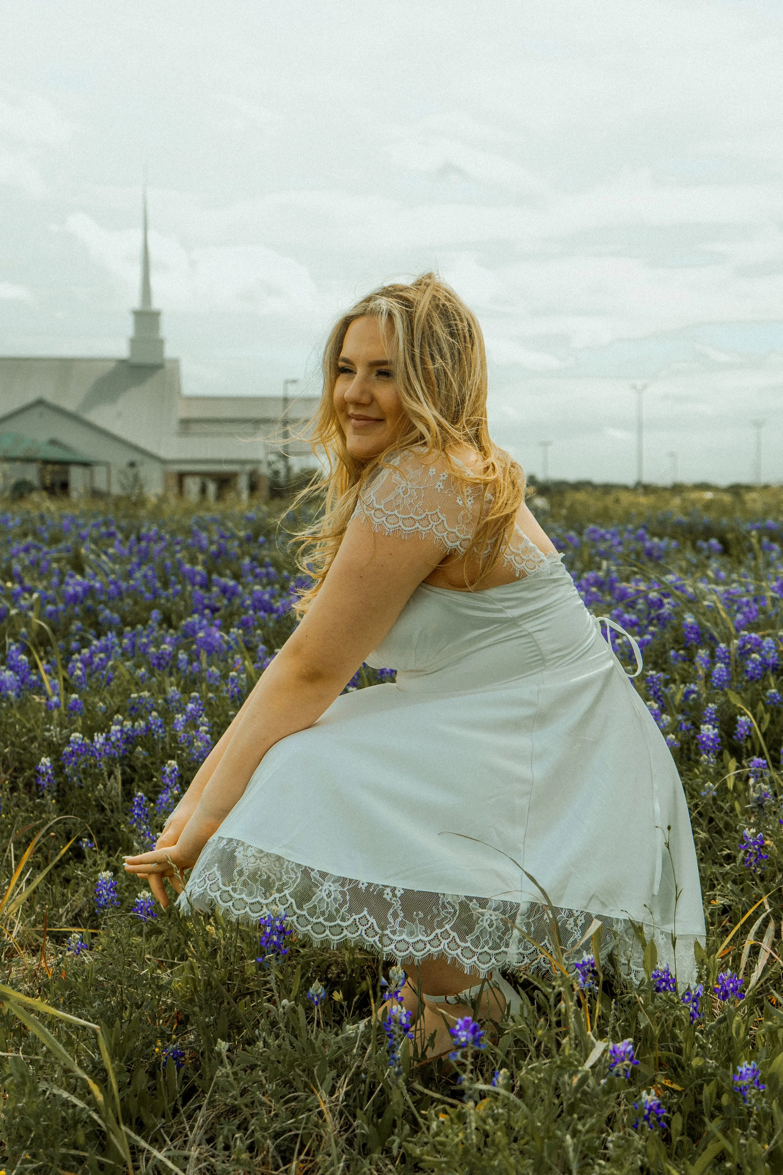 A woman in a white dress with lace details is sitting in a field of purple flowers, smiling with her eyes closed and head turned slightly to the side during daytime with cloudy sky.