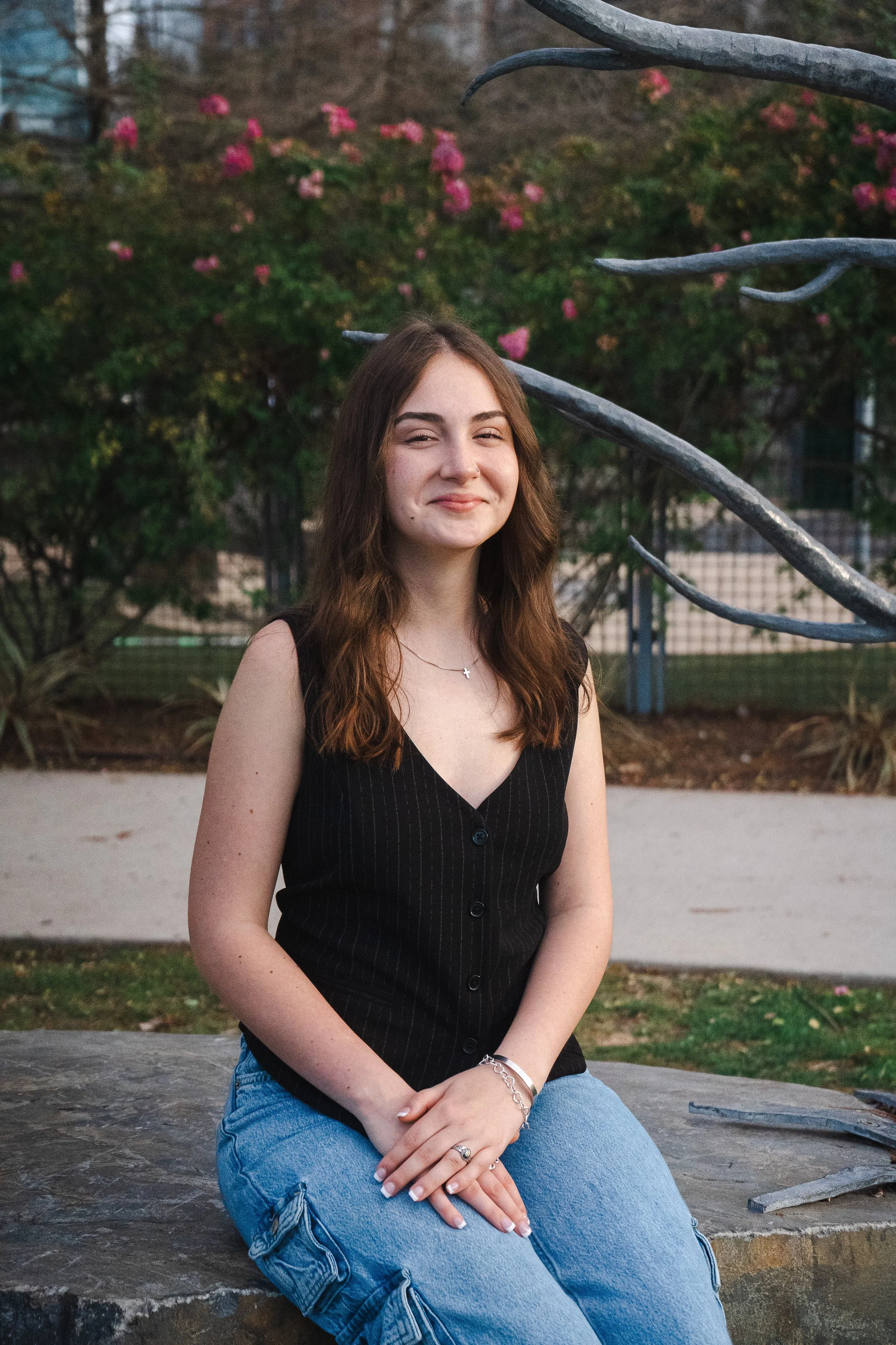 A young woman with long brown hair sitting on a stone bench outdoors, smiling, with pink flowering bushes and trees in the background.