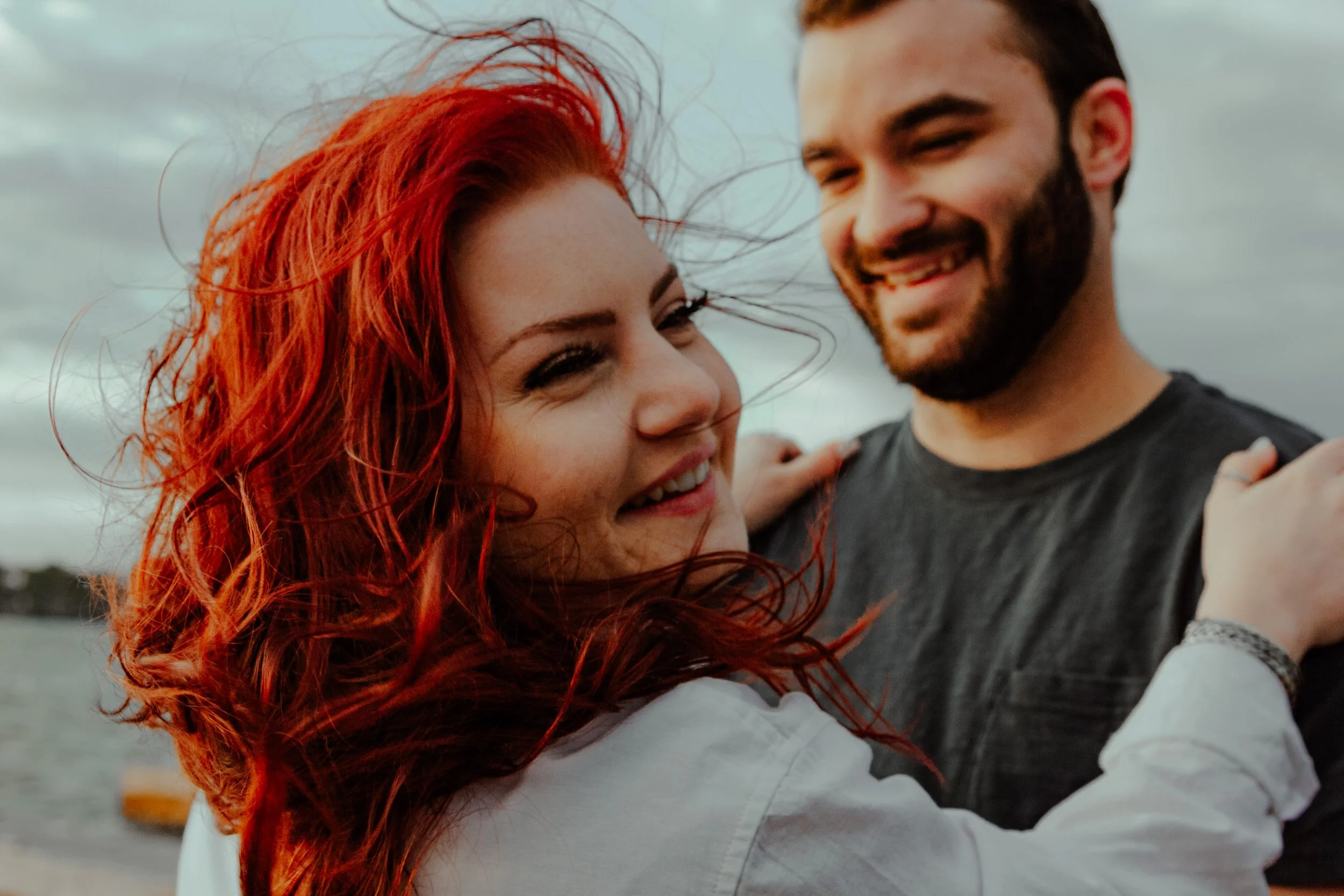 A smiling young woman with bright red curly hair embracing a bearded young man with dark hair, outdoors near water under cloudy skies.