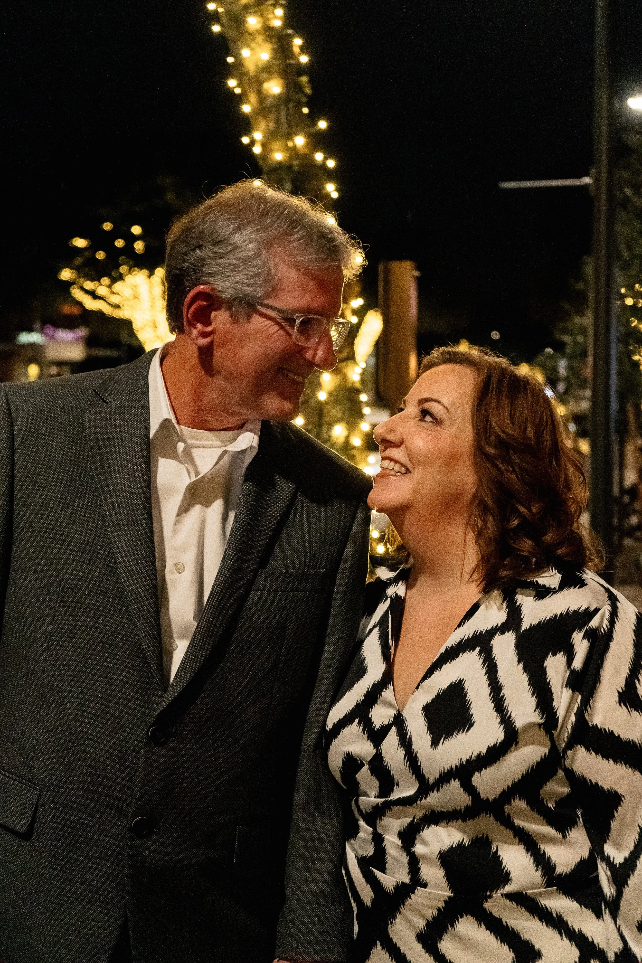 A man and woman are smiling and looking at each other, standing close together at night with decorative string lights in the background.