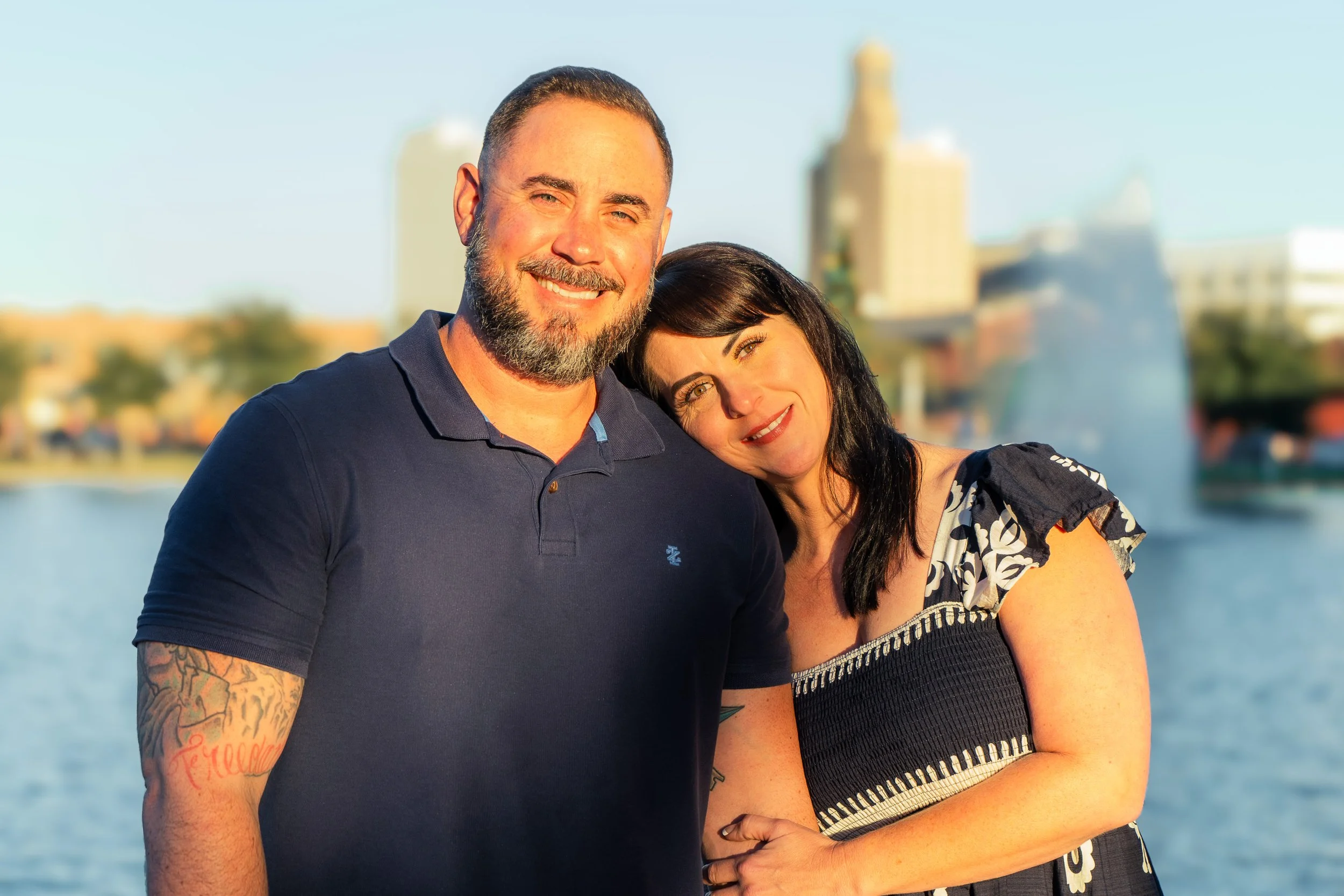A smiling man with a beard and tattoos on his arm, and a woman with black hair, in front of a river with city buildings in the background, during sunset.