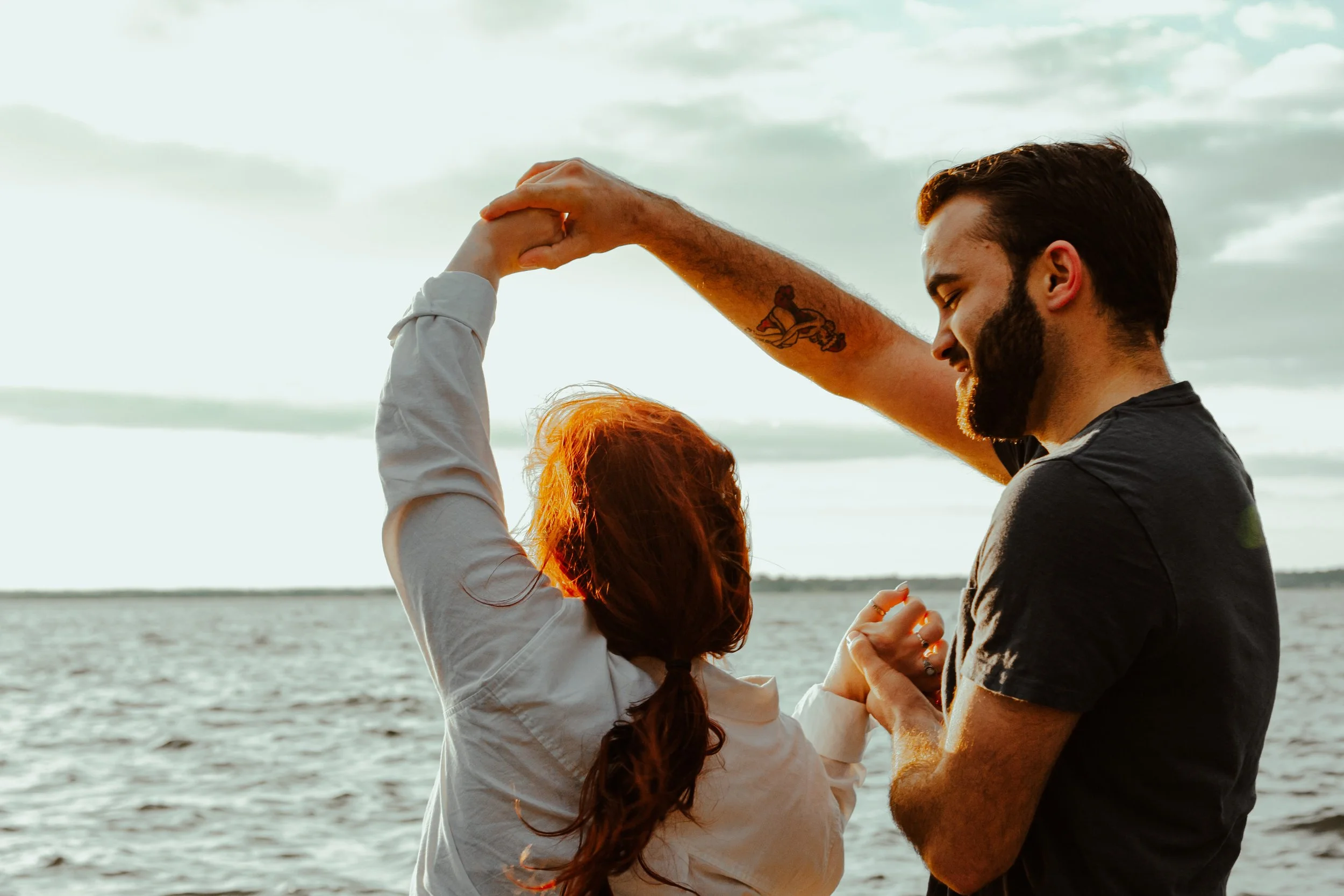 A man and woman dancing and holding hands by the water at sunset, with clouds in the sky.