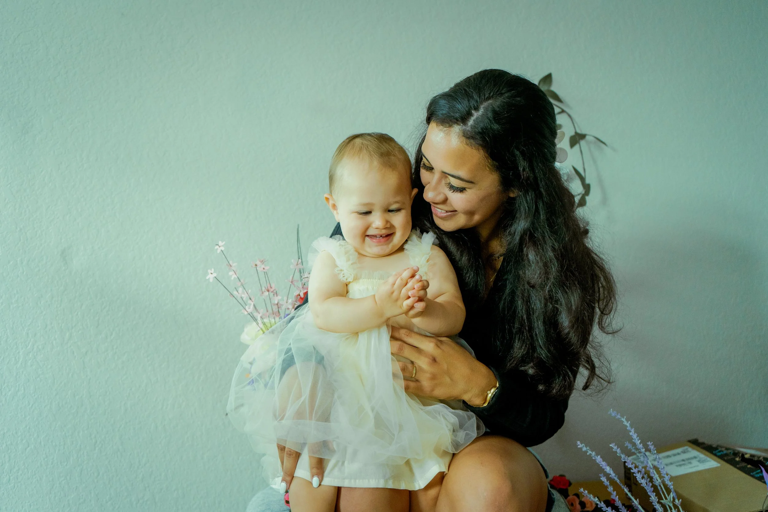 A woman with curly dark hair smiling and holding her baby daughter with blonde hair and a yellow dress, both looking happy and close together.