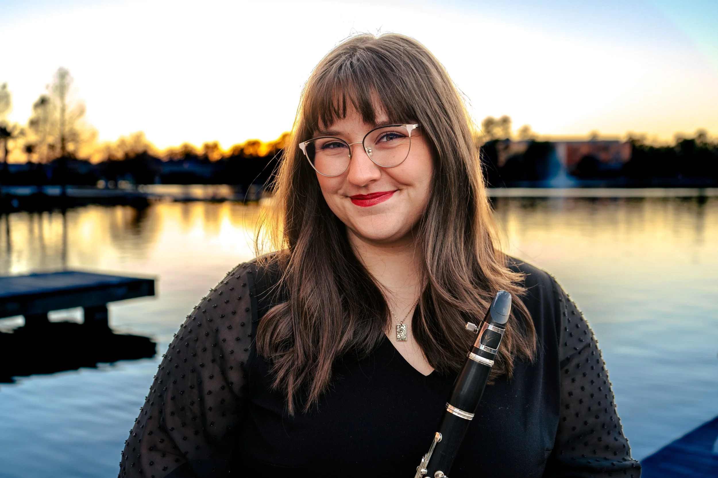 A young woman with long brown hair, glasses, and red lipstick smiling while holding a flute outdoors at sunset near a body of water with trees and buildings in the background.