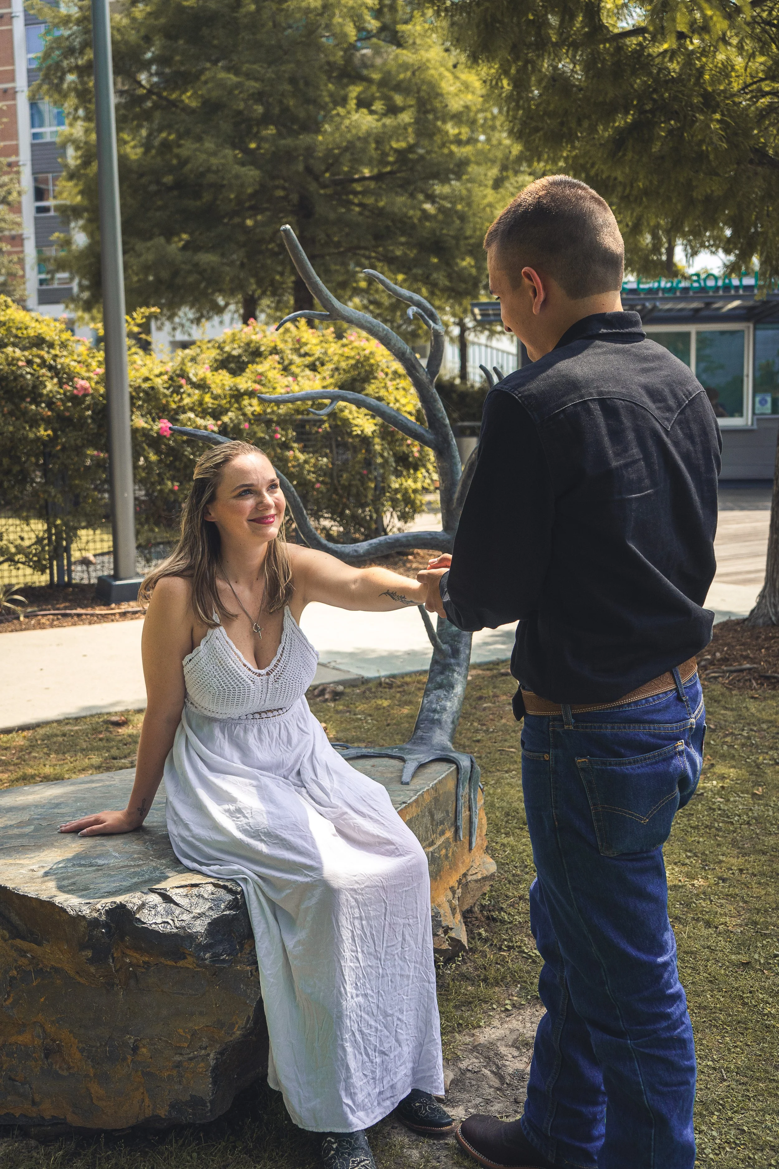 A woman in a white dress sitting on a rock, smiling and holding hands with a man in a black jacket and jeans, with a park and trees in the background.