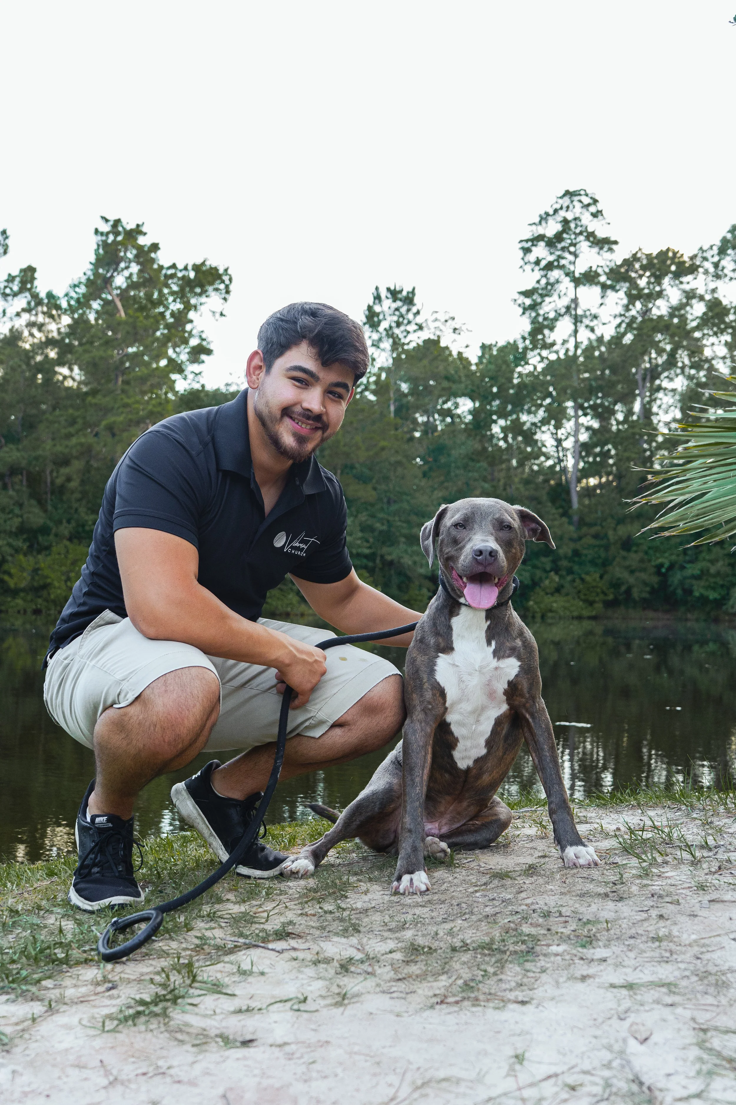 A young man crouching next to a large gray and white pitbull dog by a body of water with trees in the background.