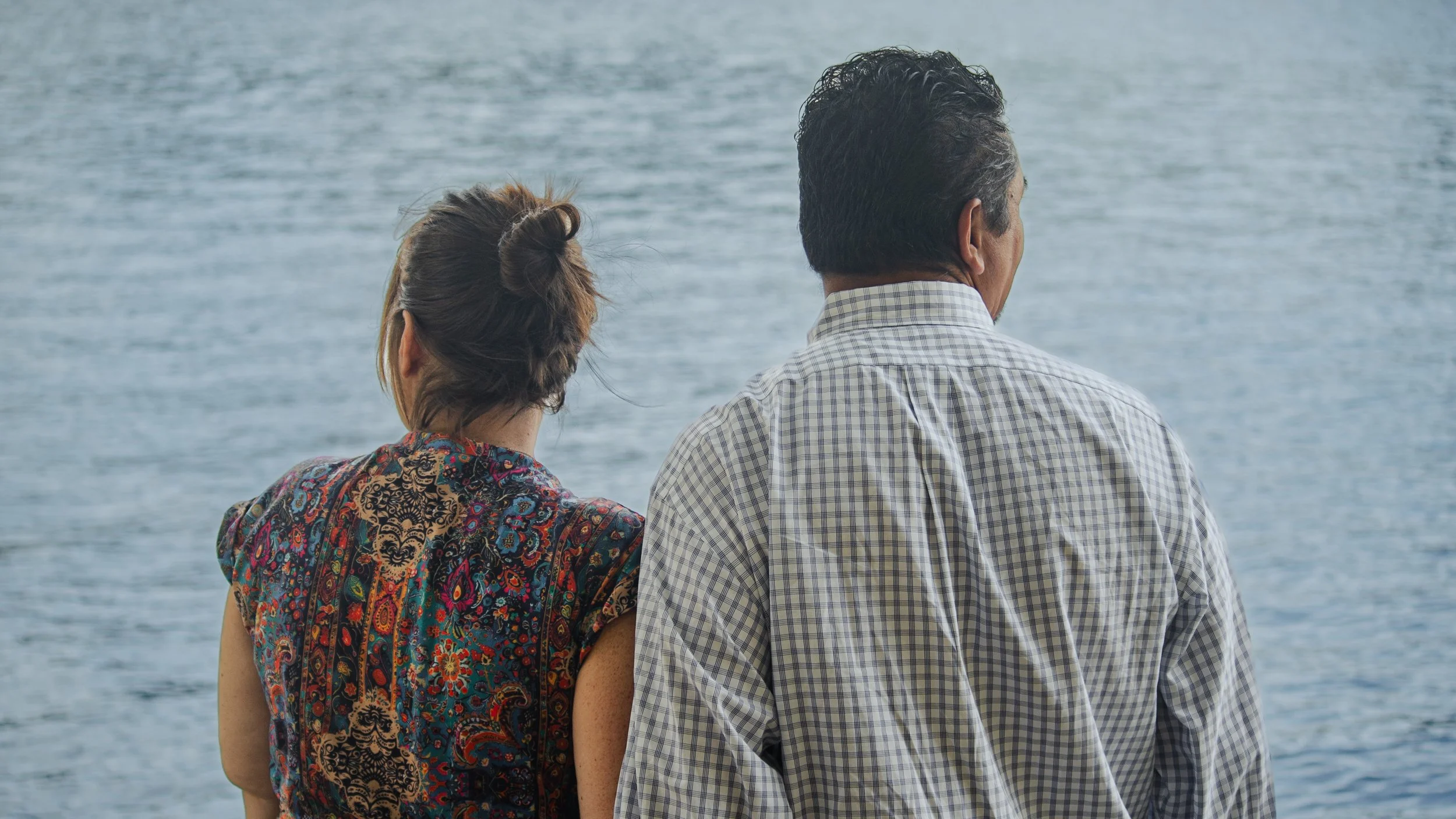 A man and woman stand together facing a body of water, with the woman's arm linked with the man's.