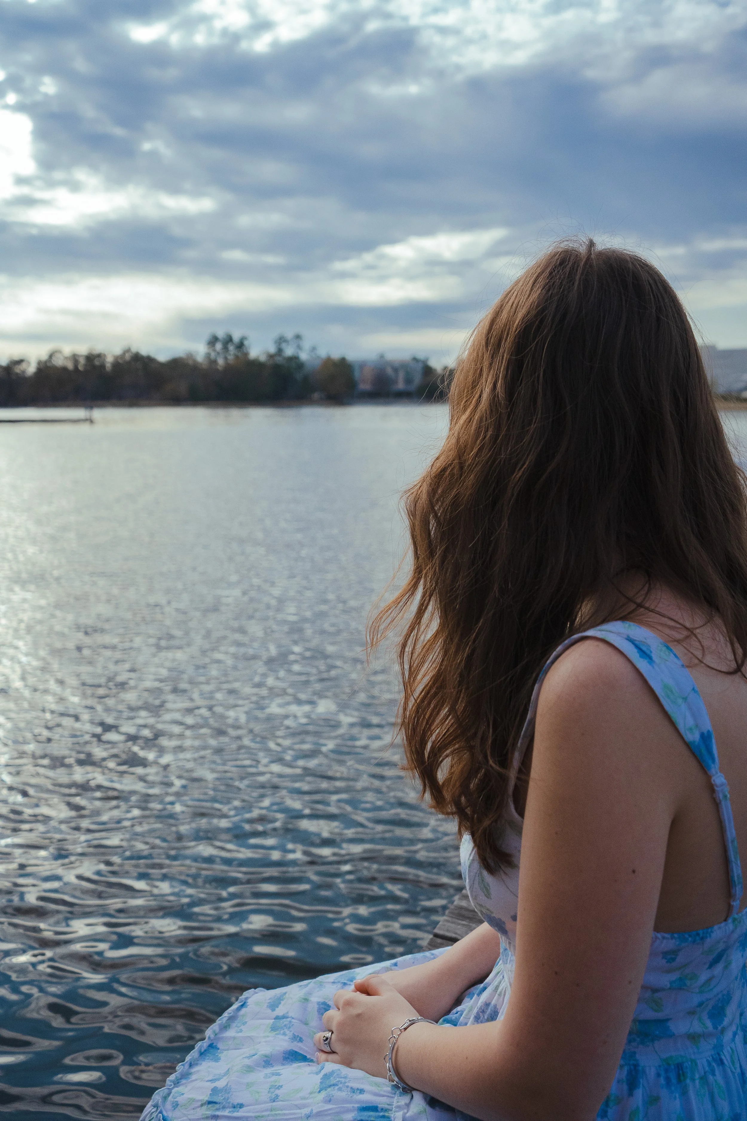 A woman with brown hair sitting by a body of water on a cloudy day, facing away from the camera, wearing a sleeveless dress with a blue and green pattern.
