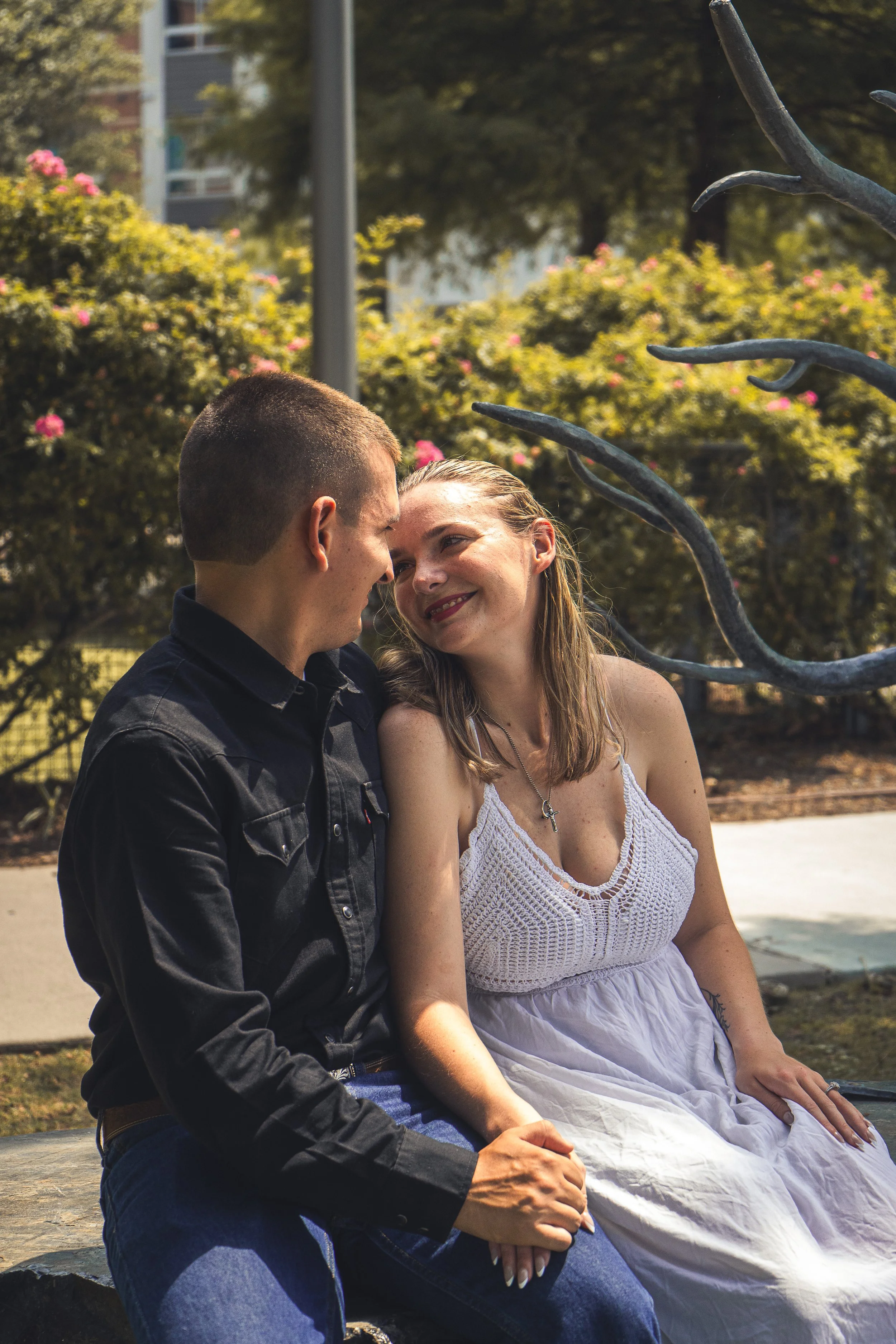 A man and a woman sitting close on a bench outdoors, touching foreheads, with trees and flowering bushes in the background.
