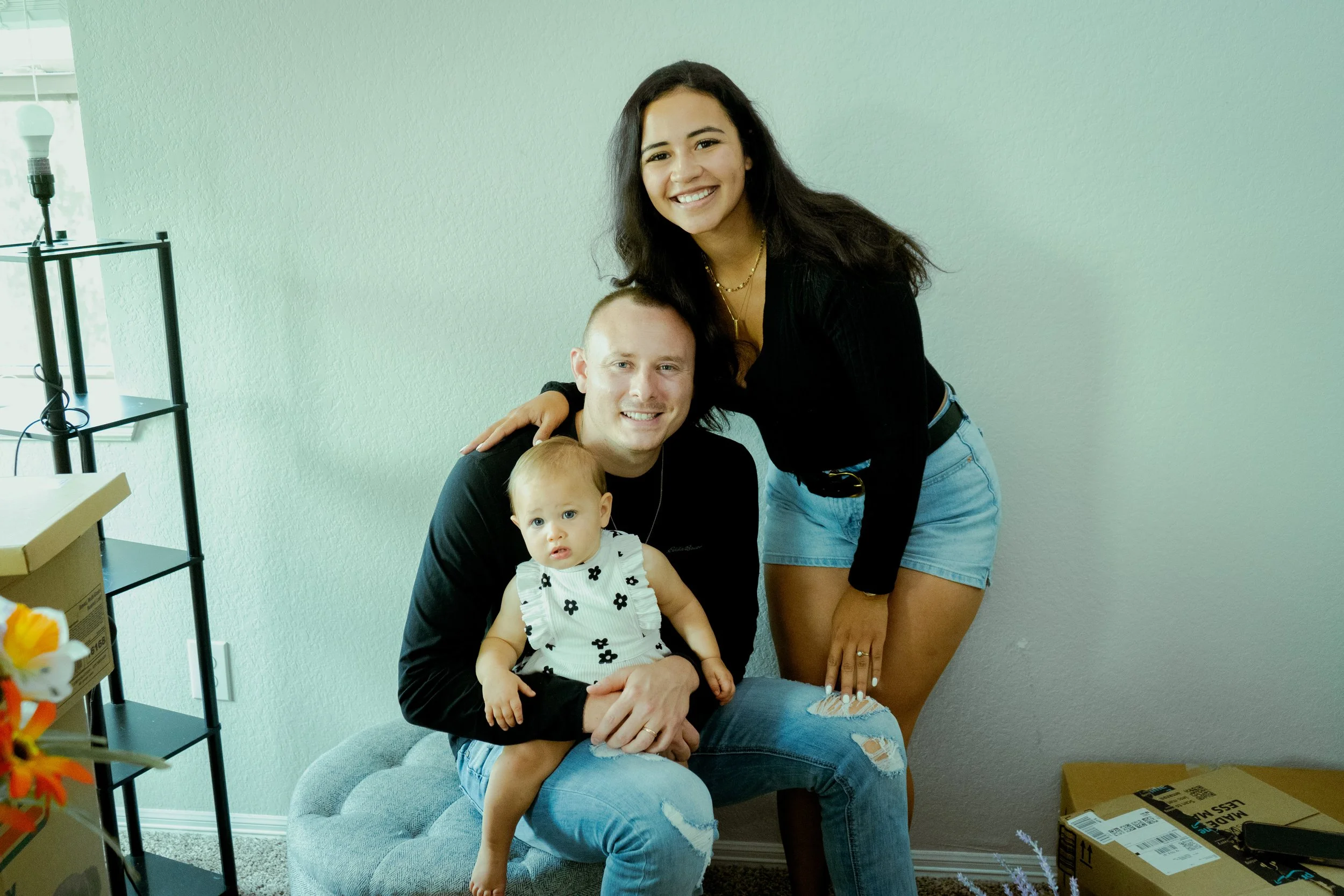 people, including a baby, pose for a photo in a room with light green walls. One woman with long dark hair, wearing a black top and denim shorts, stands behind a seated man with short blond hair, wearing a black shirt. The man holds a baby dressed in