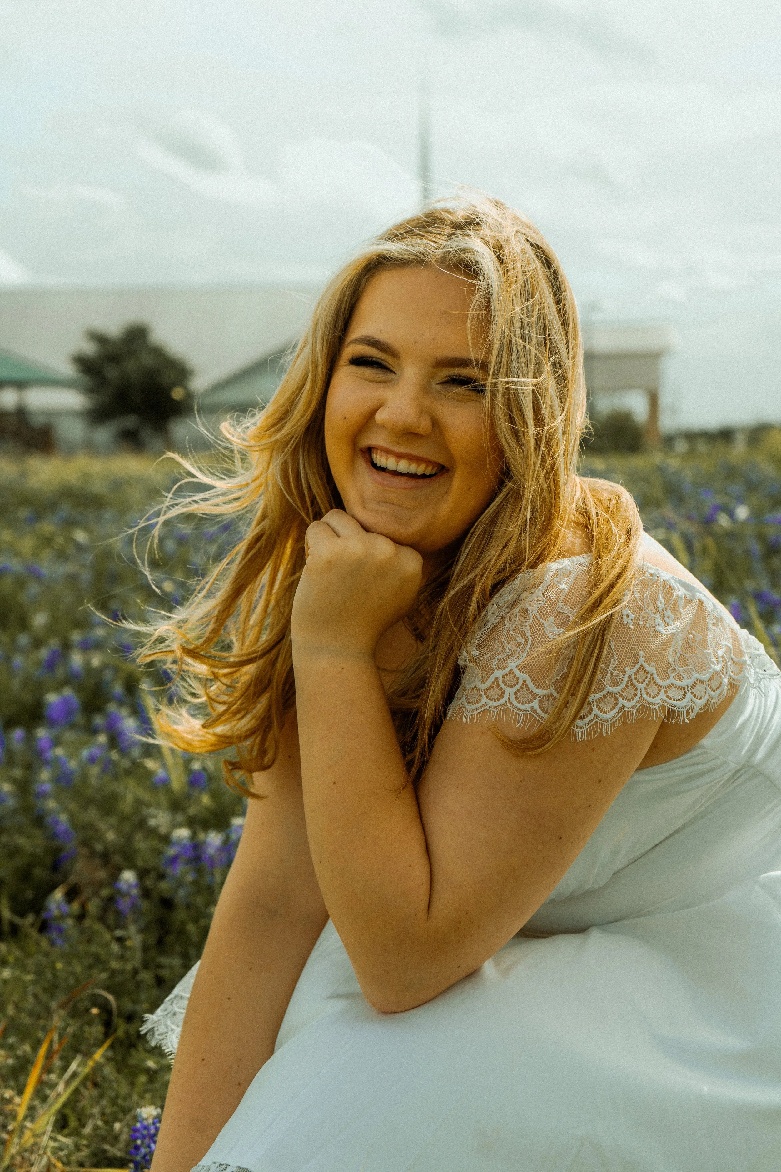 A young woman with long blonde hair sitting outdoors in a field of purple flowers, smiling and resting her chin on her hand. She is wearing a white lace dress, and the background shows a building, a tree, and a cloudy sky.