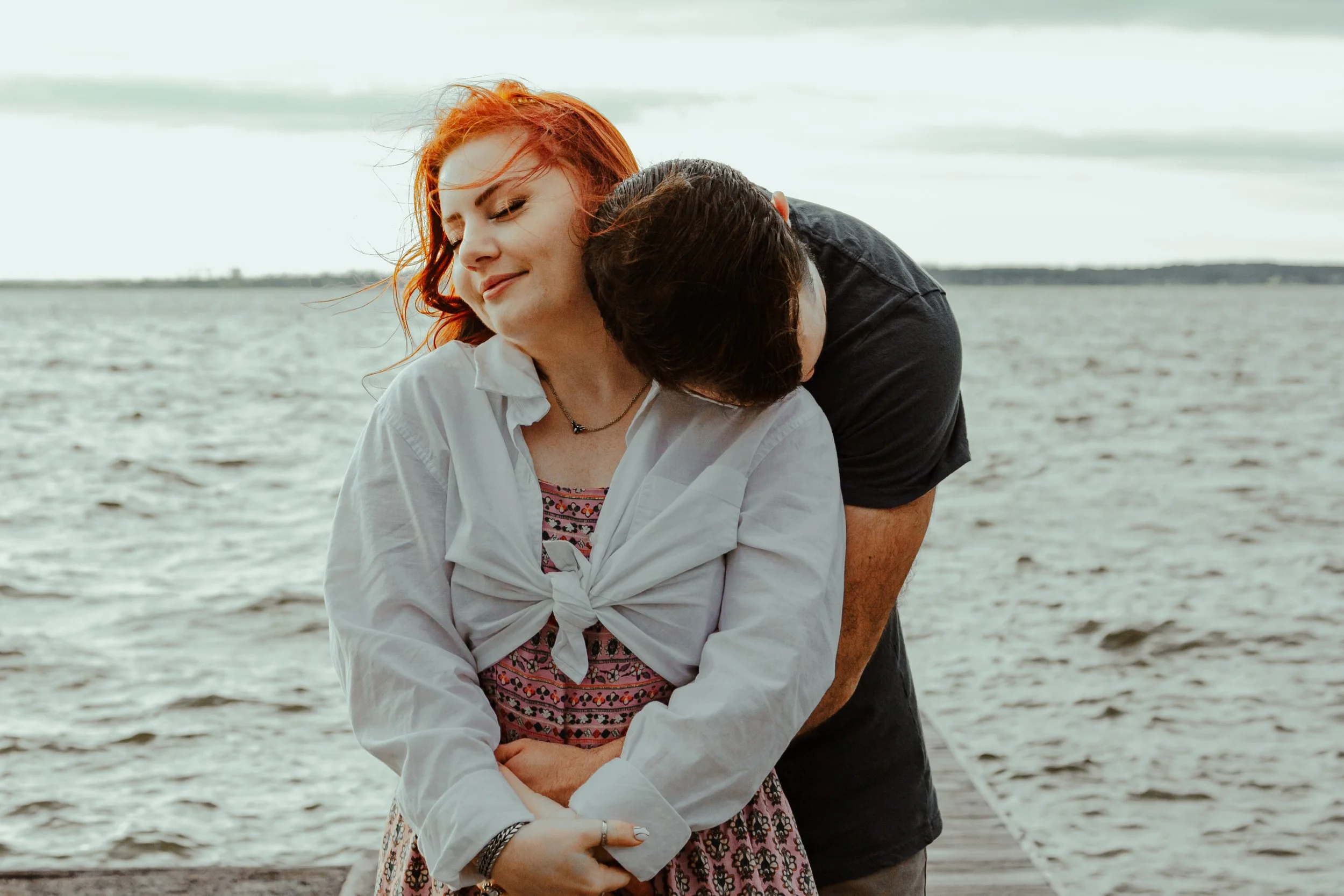 A young woman with red hair smiling with her eyes closed, being embraced from behind by a person whose face is not visible. They are by a body of water with a cloudy sky above.