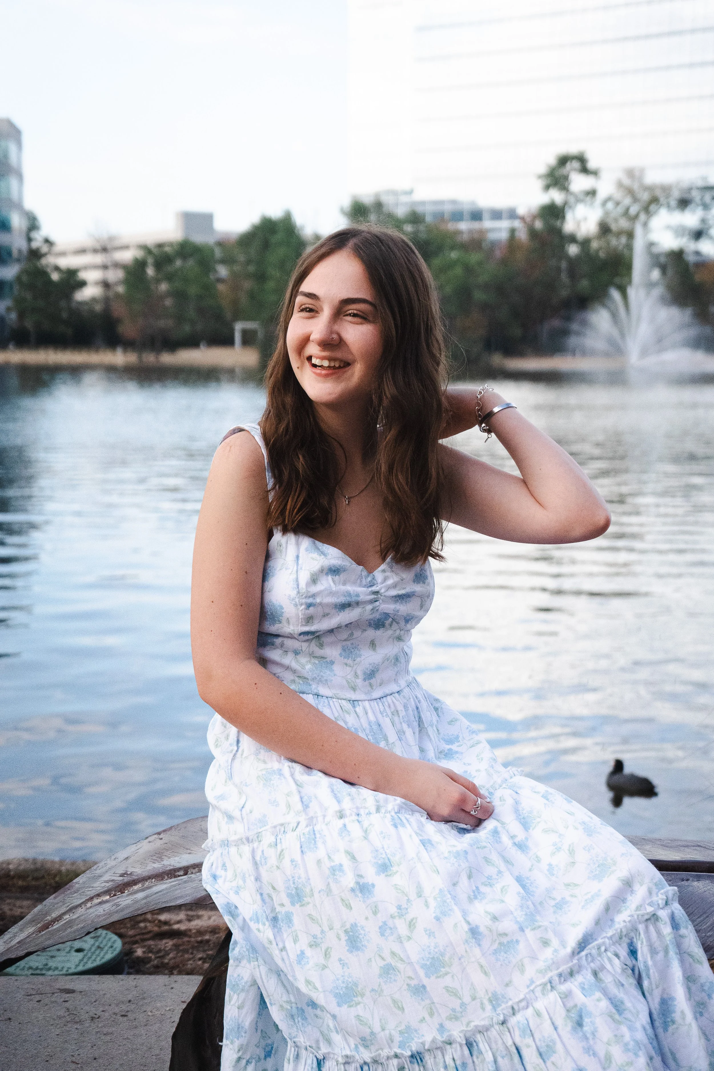 A young woman with brown hair, wearing a white dress with light blue and green floral patterns, sitting on a large log near a body of water with a fountain in the background, smiling and looking to her left.