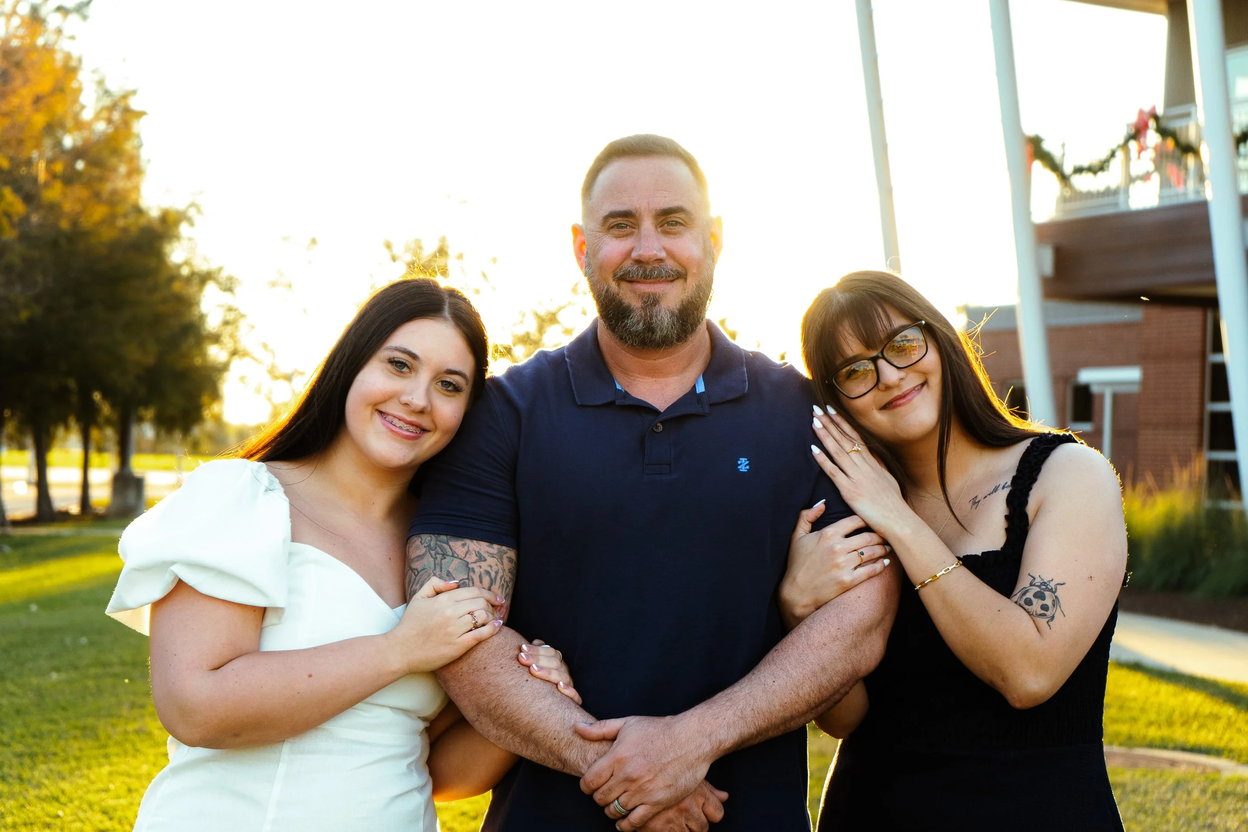 A smiling man with a beard and tattoos, flanked by his two young daughters, one with glasses, in an outdoor park setting during sunset.