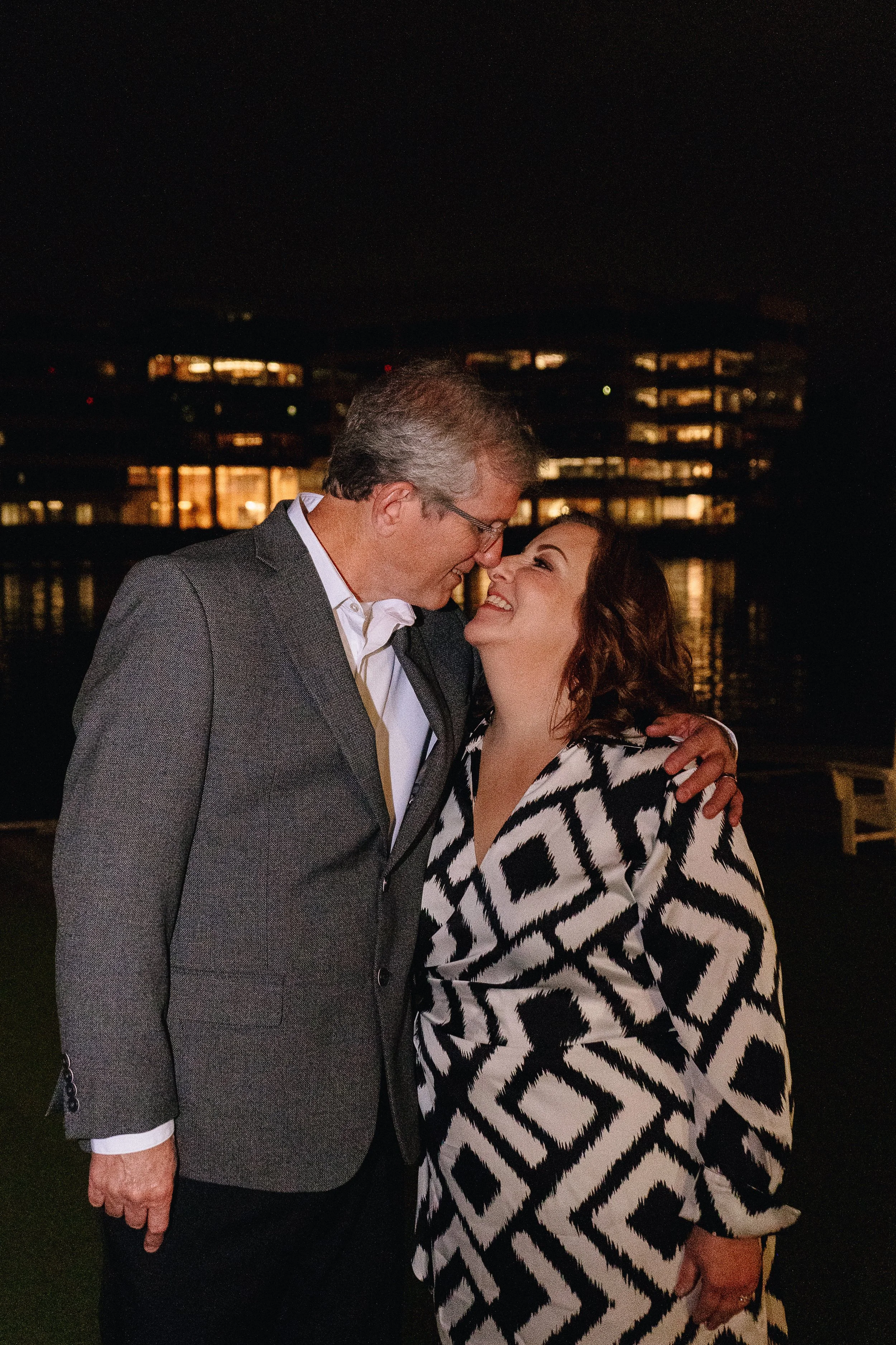 A man and a woman are smiling and touching foreheads at night with a cityscape and water in the background.