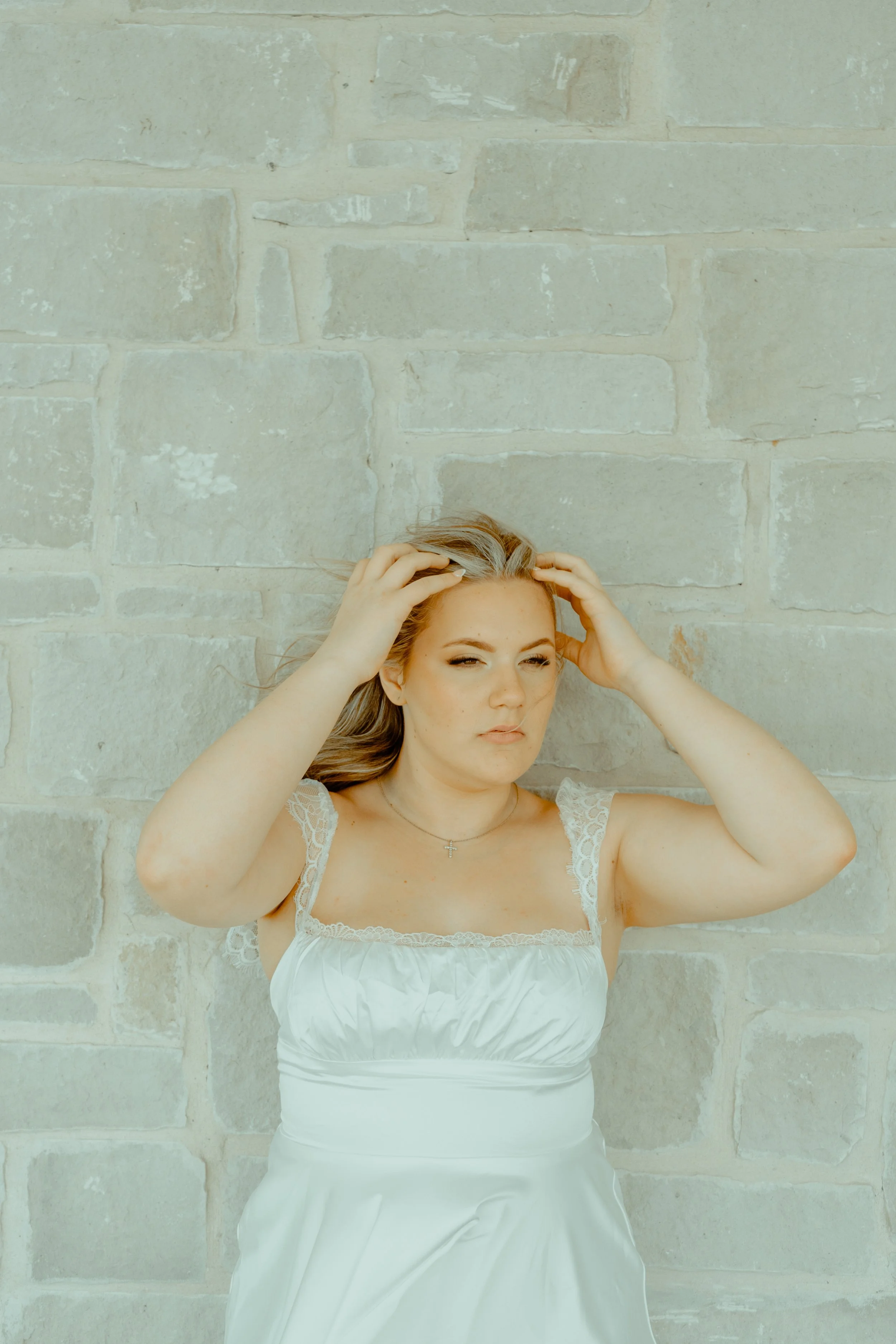 A young woman with long, light brown hair touches her head with both hands and is posing against a light-colored stone brick wall. She is wearing a white top with lace details and a white skirt, and has a small cross necklace. She has a confident fac