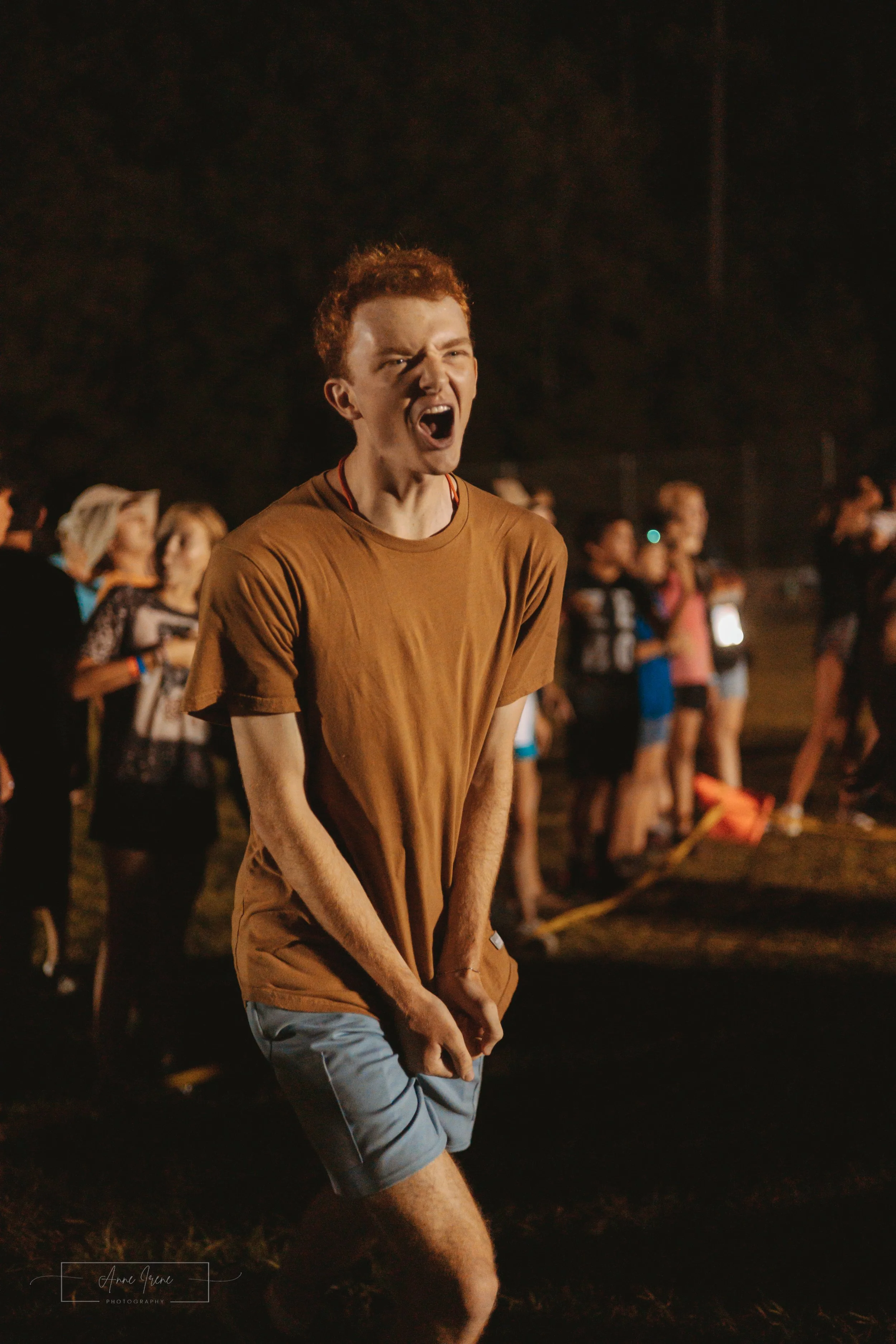 A young man with red curly hair yelling at a church camp night time game. He is wearing a brown T-shirt and gray shorts, with people and tents visible in the background.