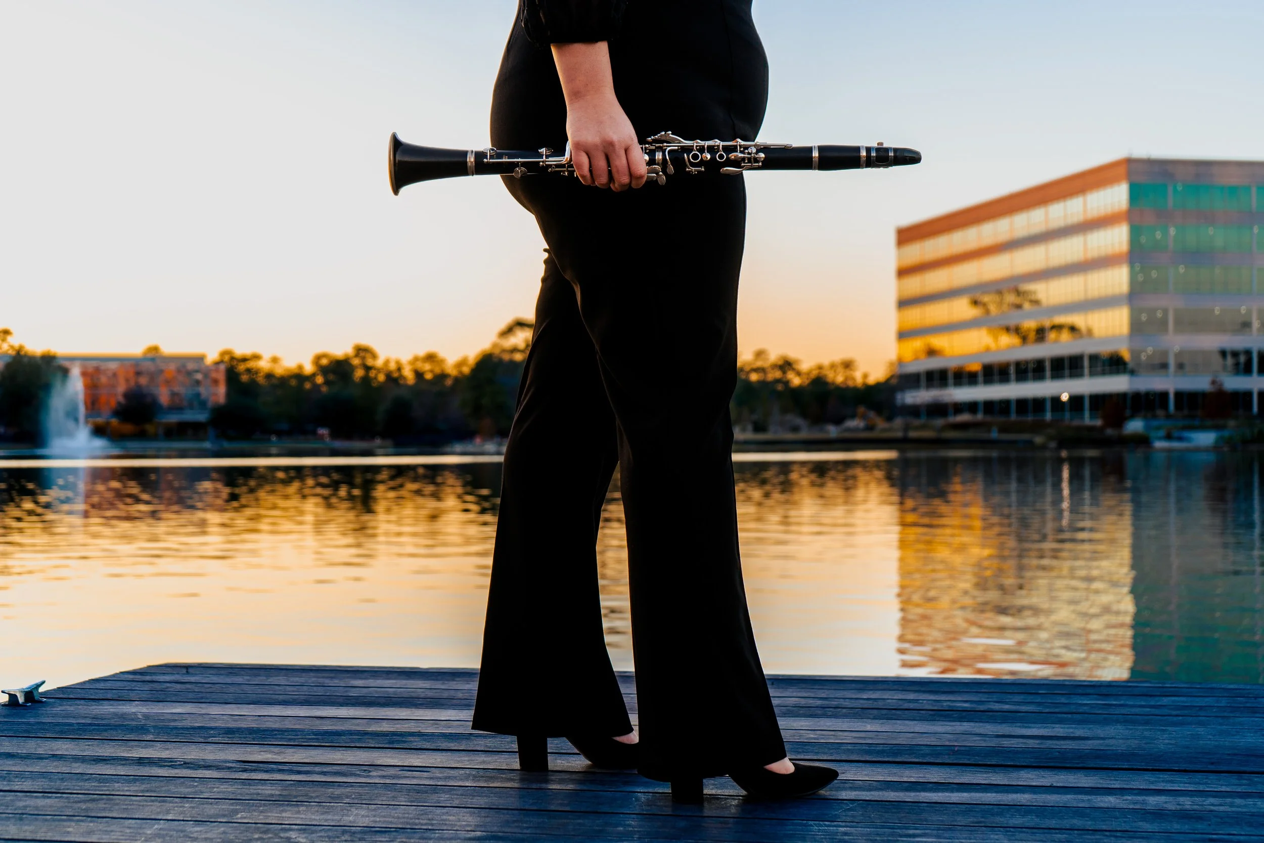 A young woman standing on a dock holding a clarinet in their right hand, with a body of water, trees, and a modern multicolored building in the background during sunset.
