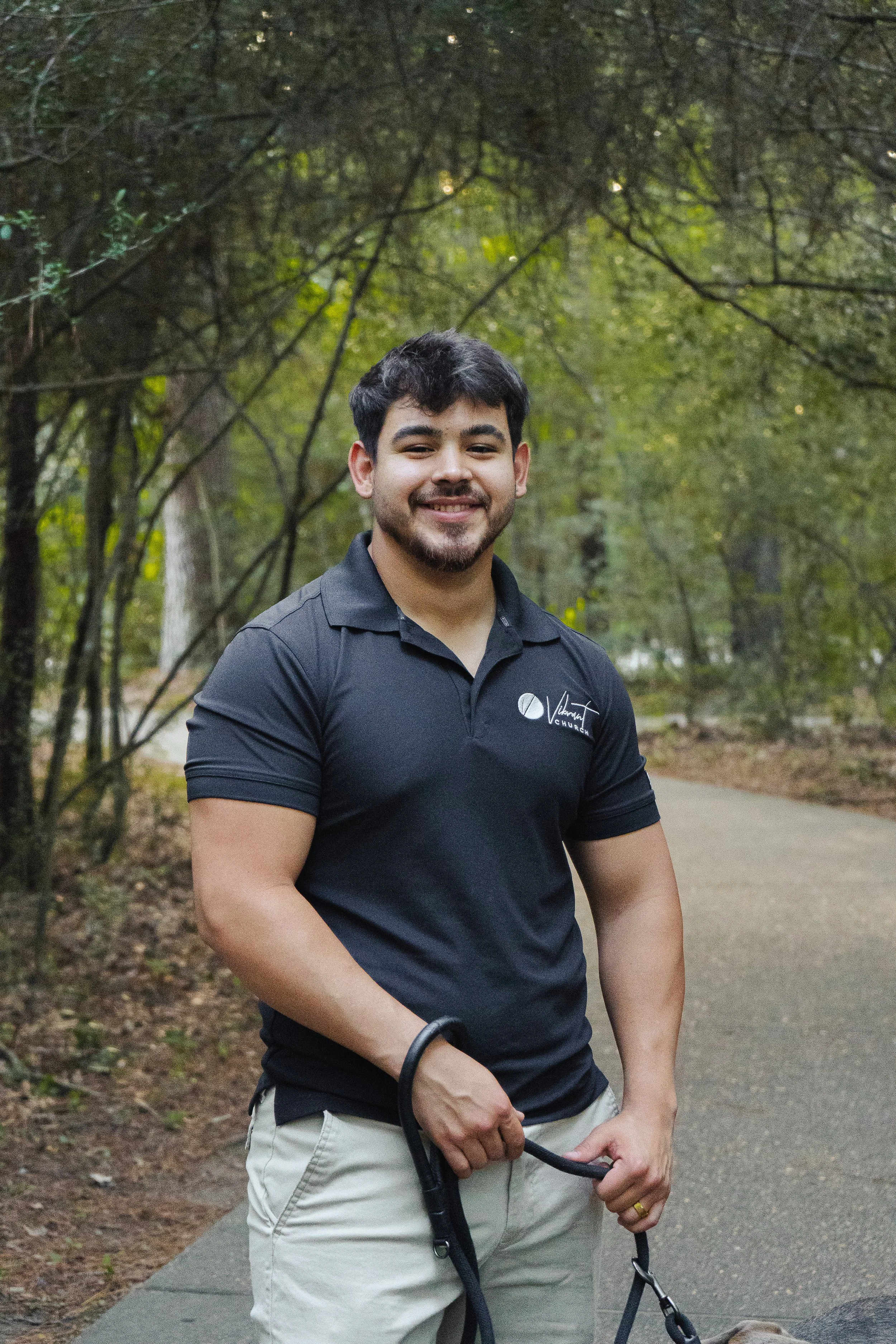 A young man with dark hair and a beard, smiling, dressed in a black polo shirt and light-colored shorts, standing on a paved path in a wooded park, holding the leash of a dog that is partially visible at the bottom of the image.