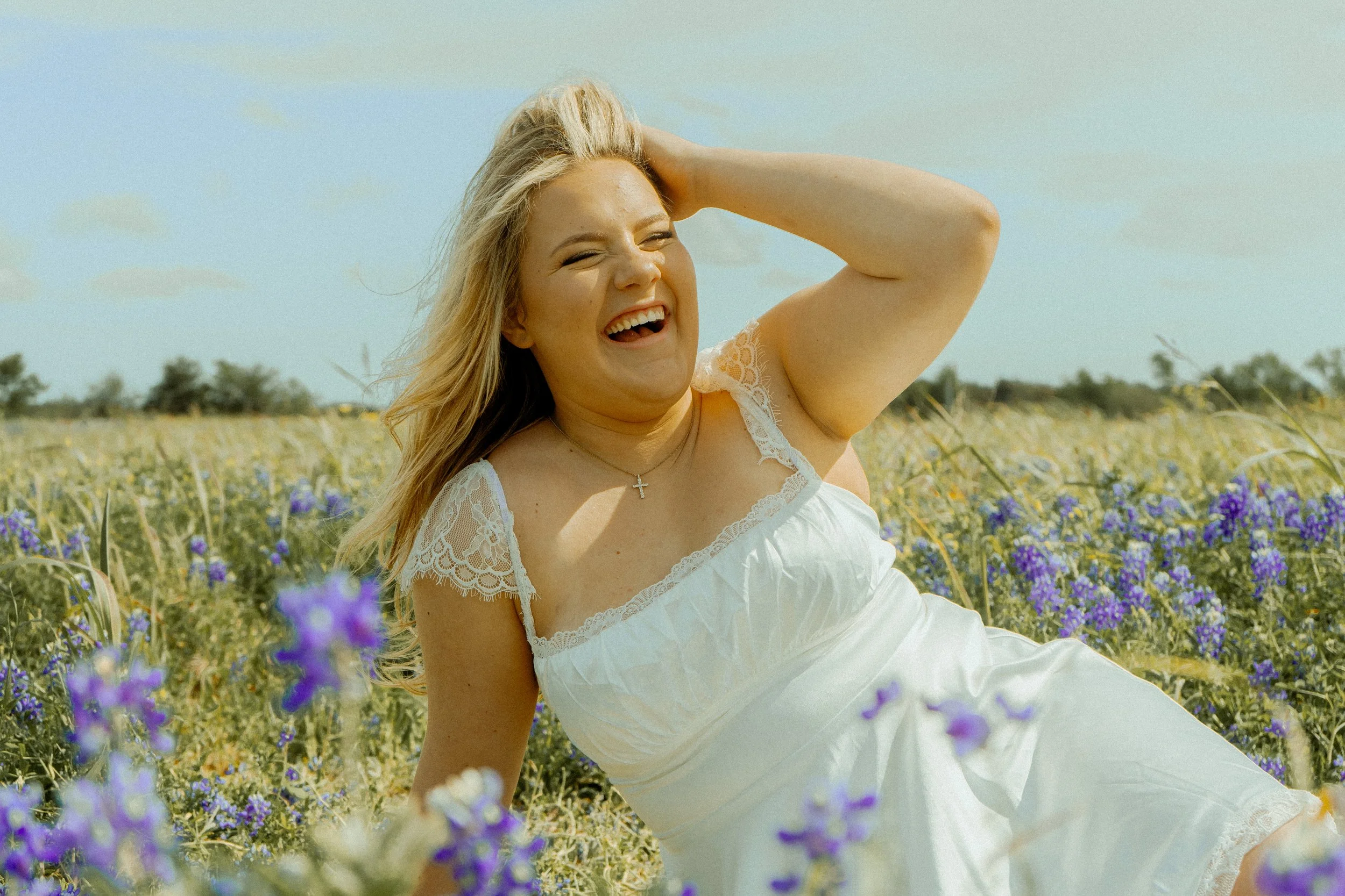 A woman with blonde hair smiling and holding her head with one hand in a field of purple flowers on a sunny day.