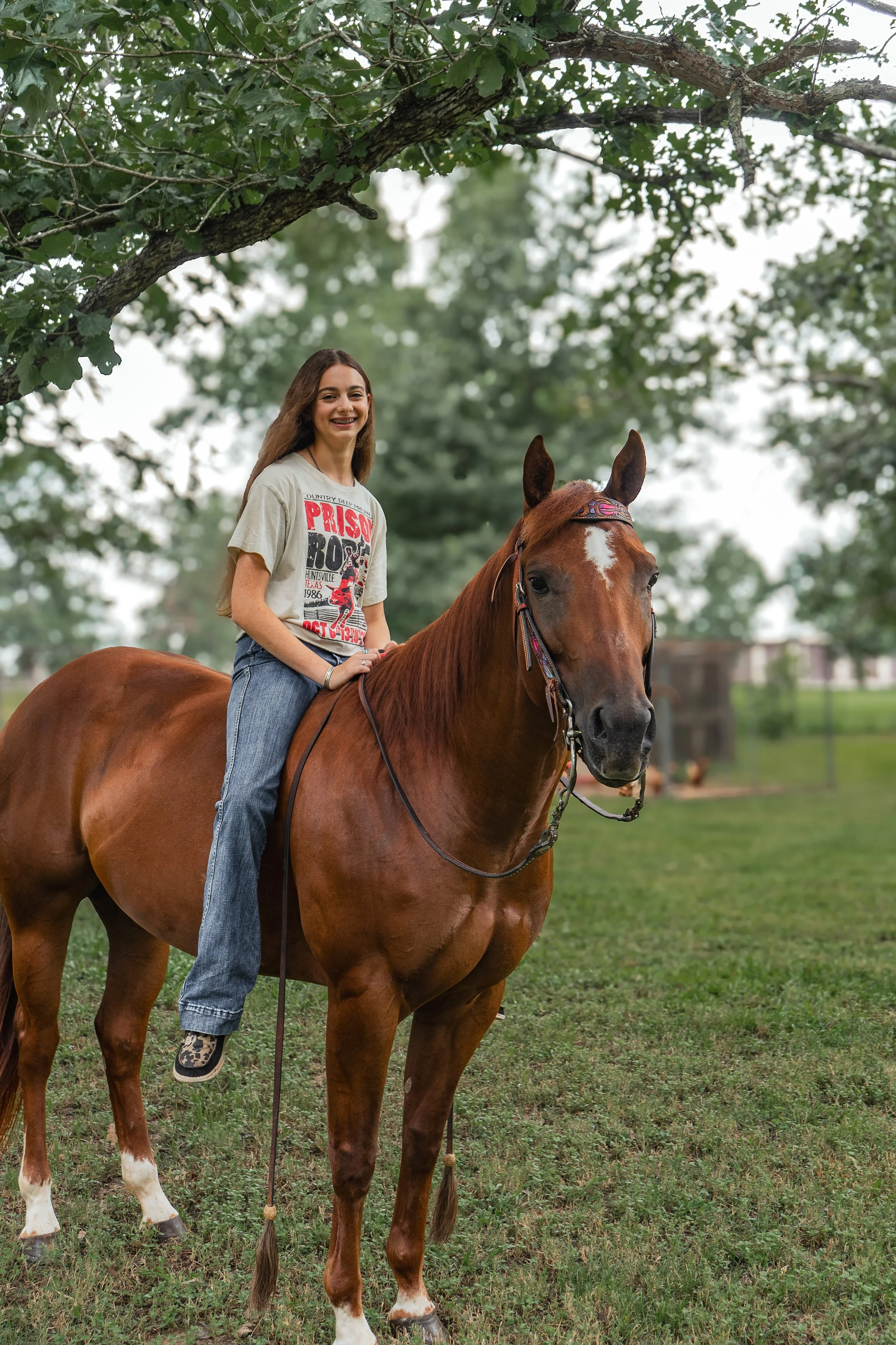 A young woman with long brown hair riding a brown horse in a park with green trees in the background. She is smiling and wearing a beige T-shirt with red, black, and white graphic text, blue jeans, and leopard print shoes.