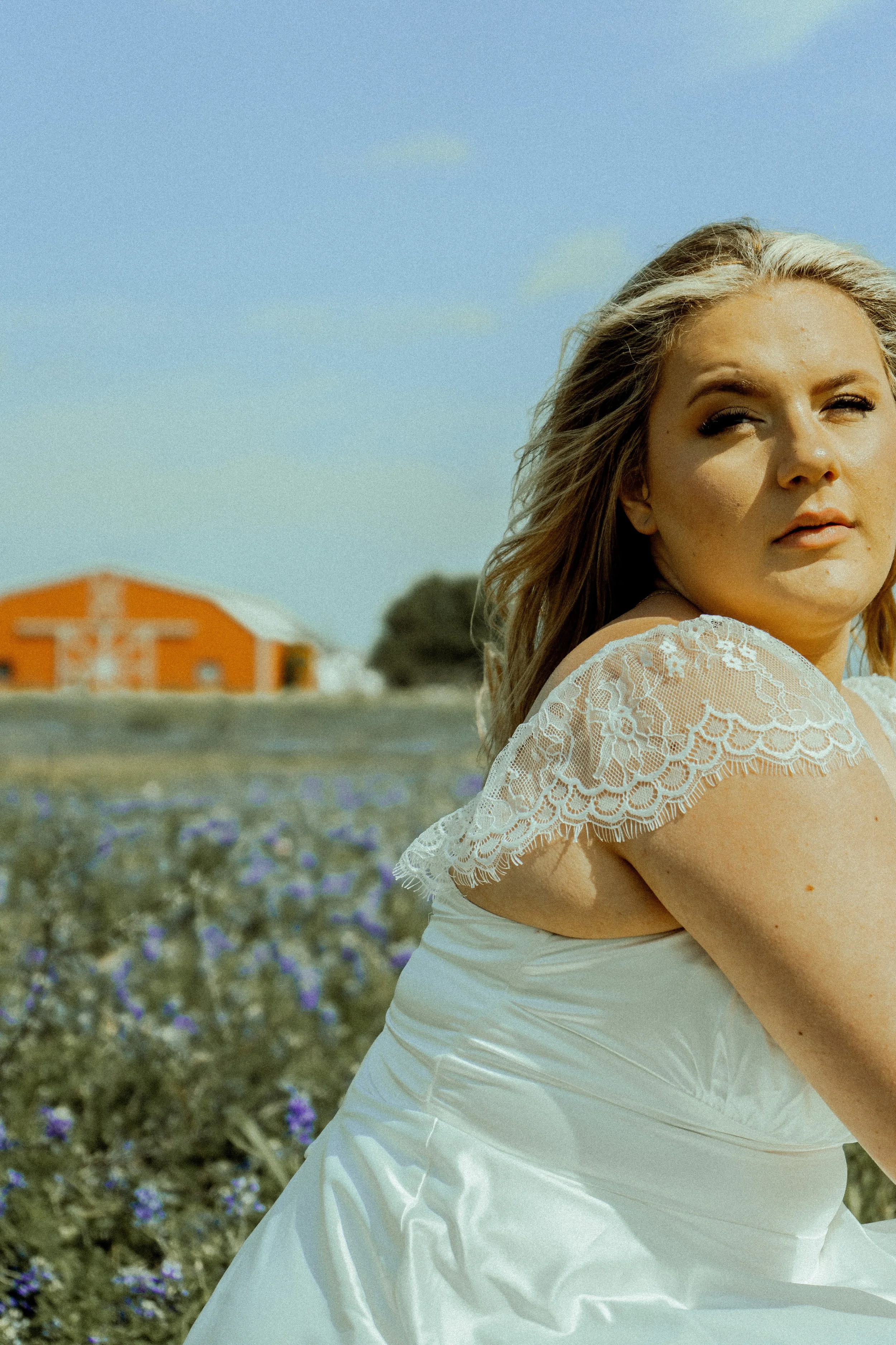A woman with blonde hair in a white dress with lace shoulder details standing in a field of purple flowers with a barn in the background.