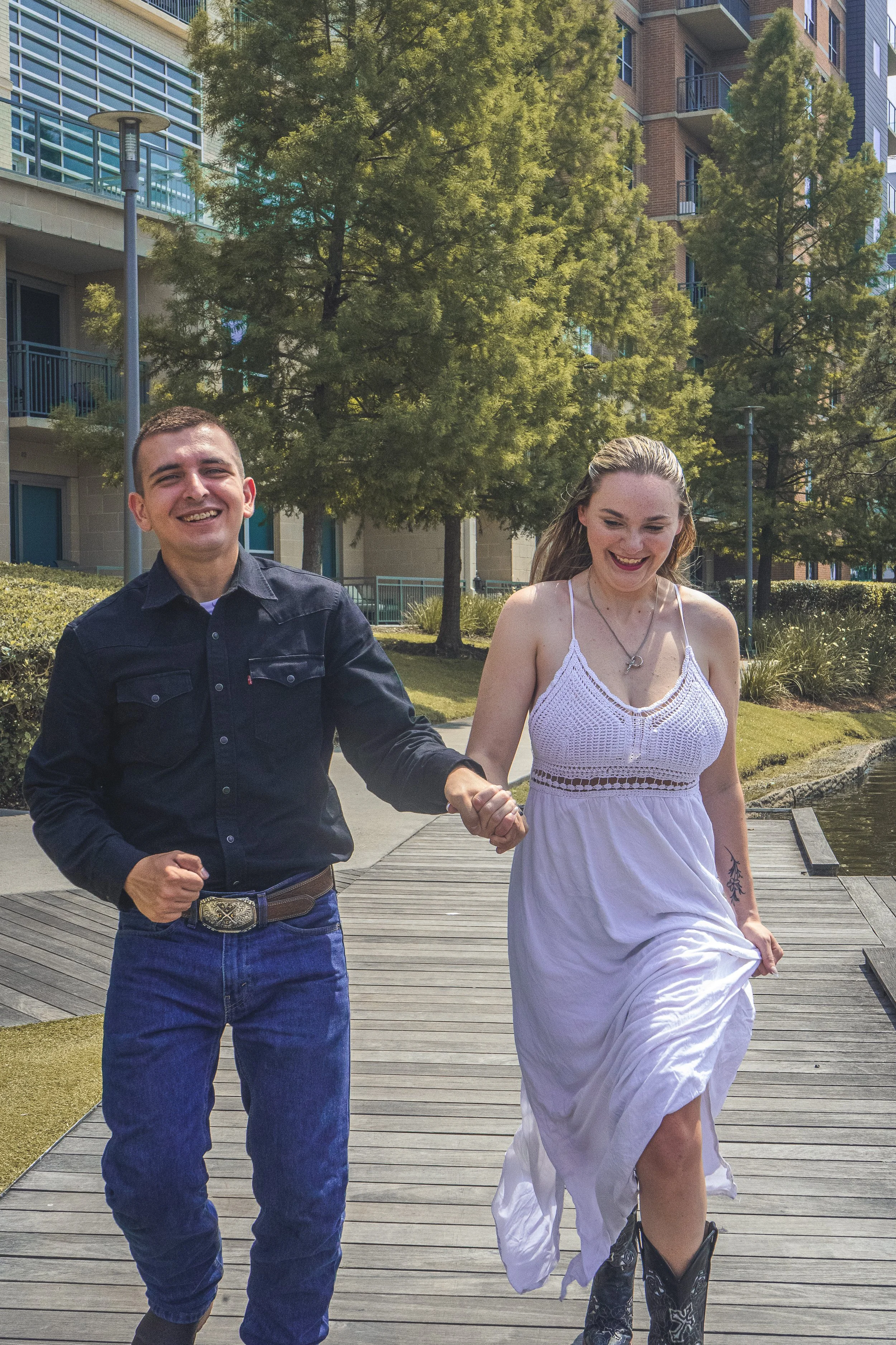 A man and woman holding hands, walking outdoors on a wooden pathway with buildings and green trees in the background.