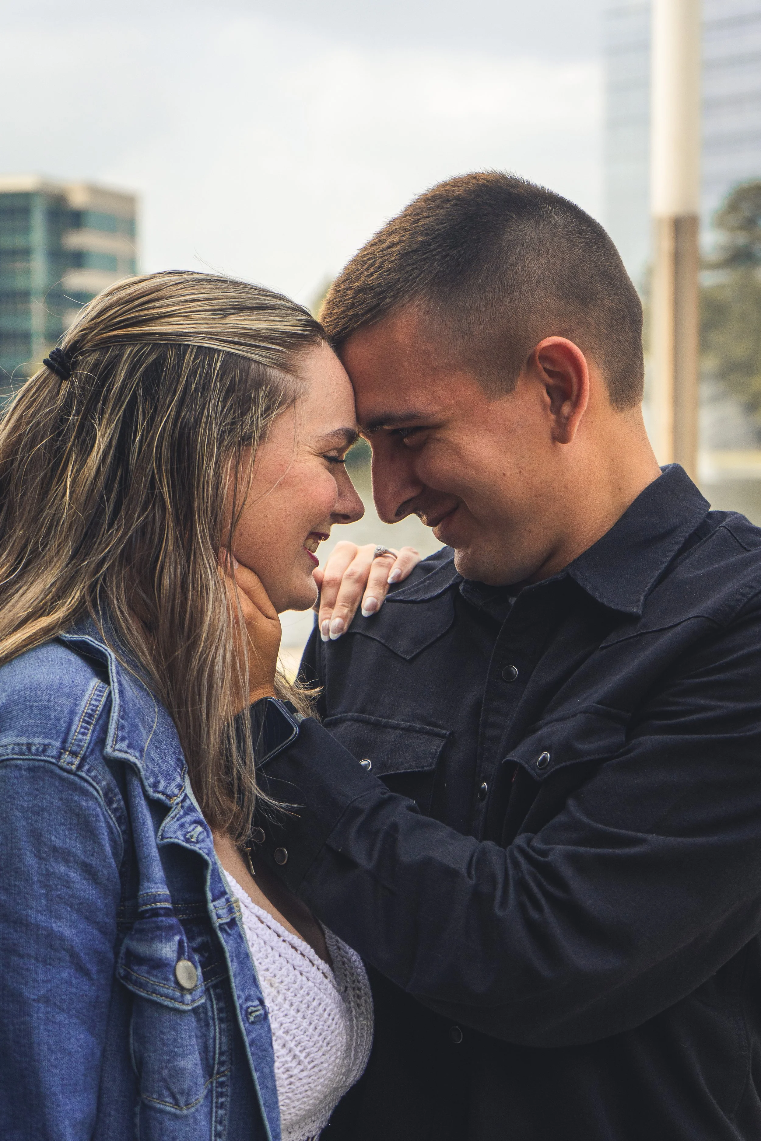 A couple with their foreheads touching, smiling, and holding each other's face, with an urban background of buildings and windows.