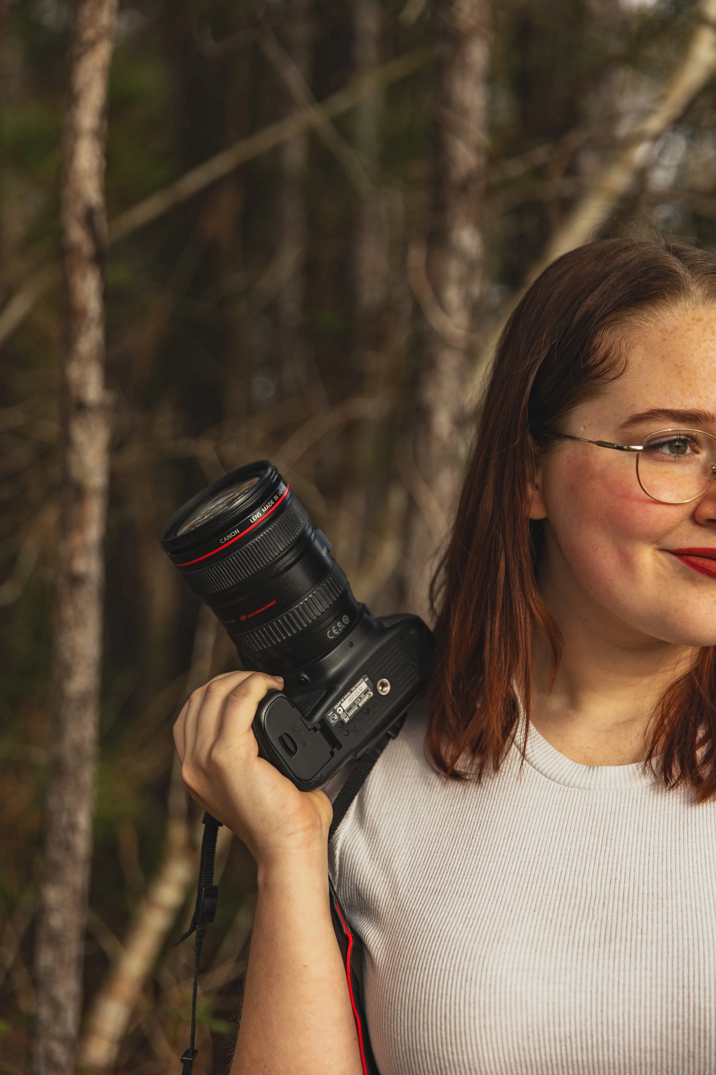 Close-up of a woman with red hair and glasses, holding a professional camera with a large telephoto lens, outdoors in a wooded area.