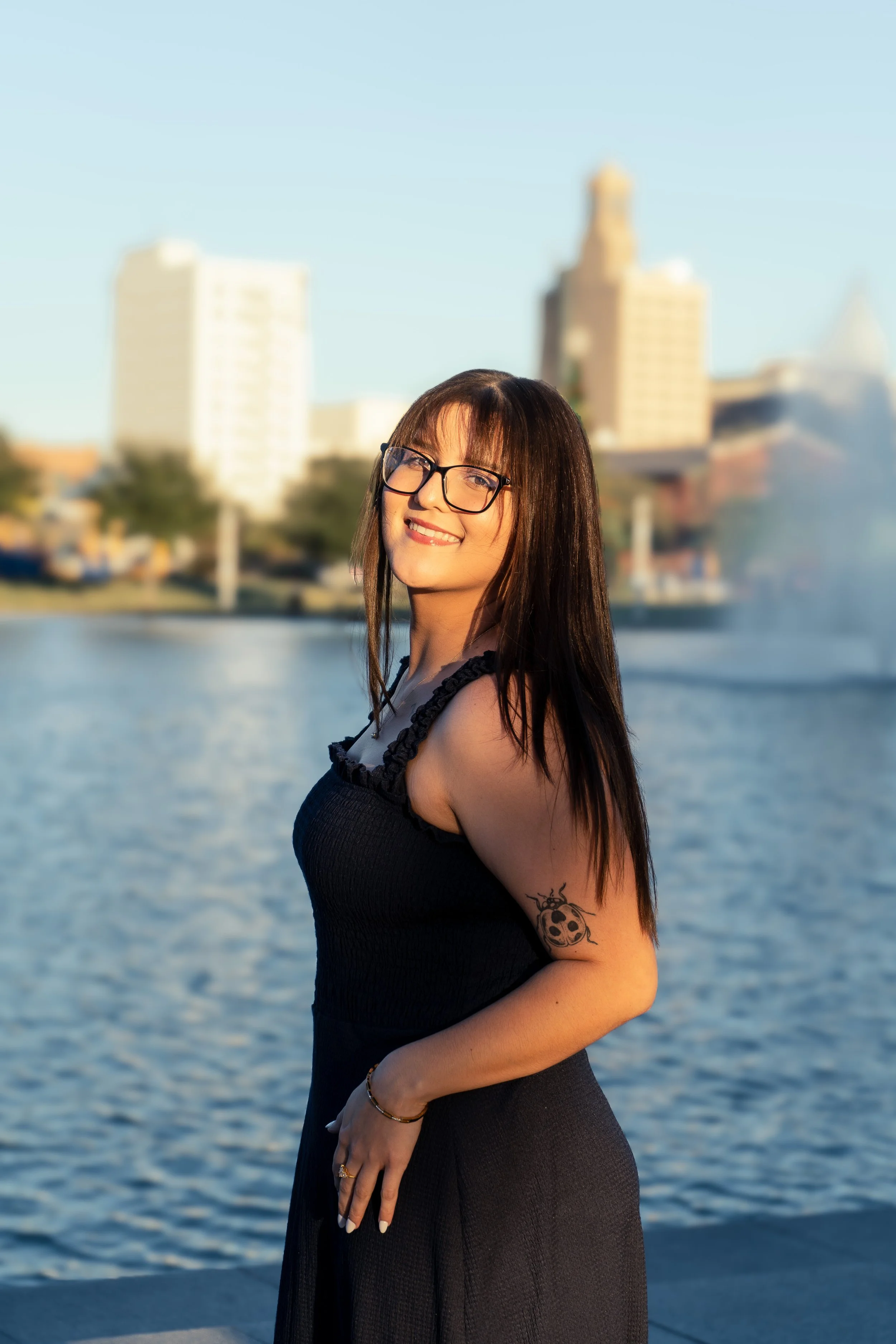 A smiling woman with long dark hair and glasses, wearing a black dress with lace trim, standing by a body of water with a city skyline in the background.