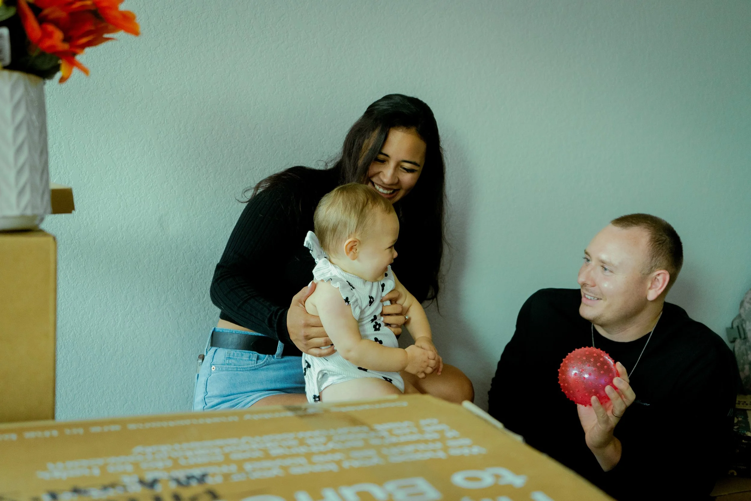 A woman smiling and holding a toddler girl during a celebration, with a man sitting nearby holding two red spiky balloons.