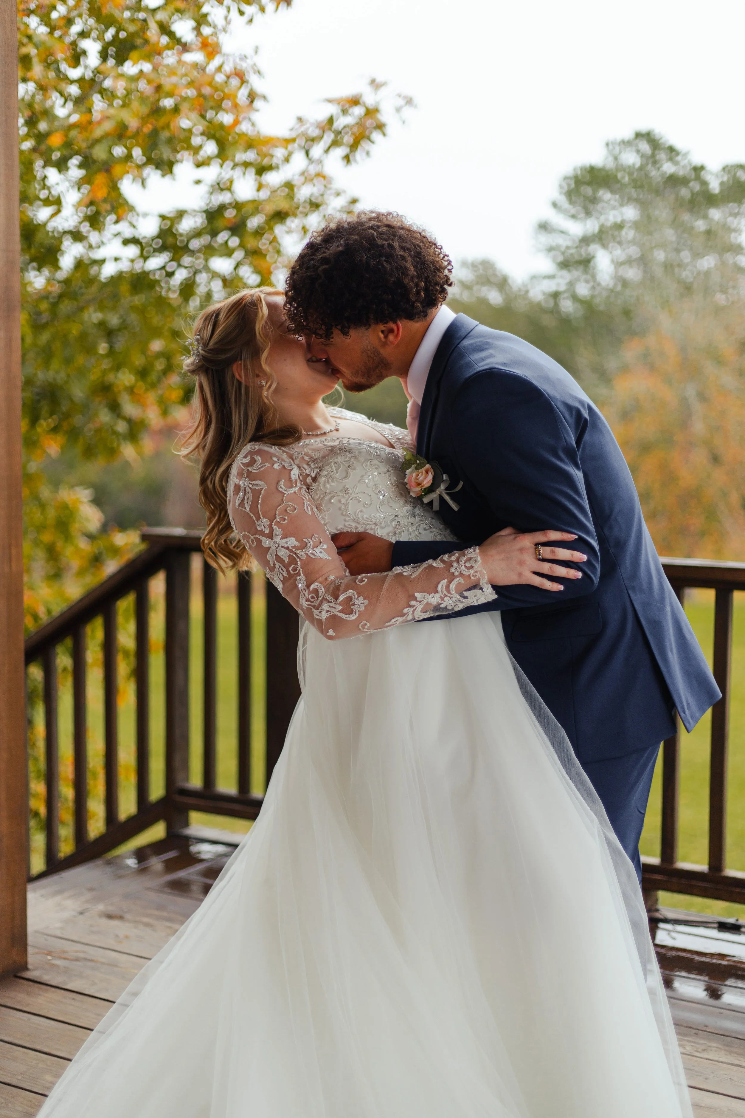 A bride and groom sharing a kiss on an outdoor wooden deck, with autumn trees in the background.