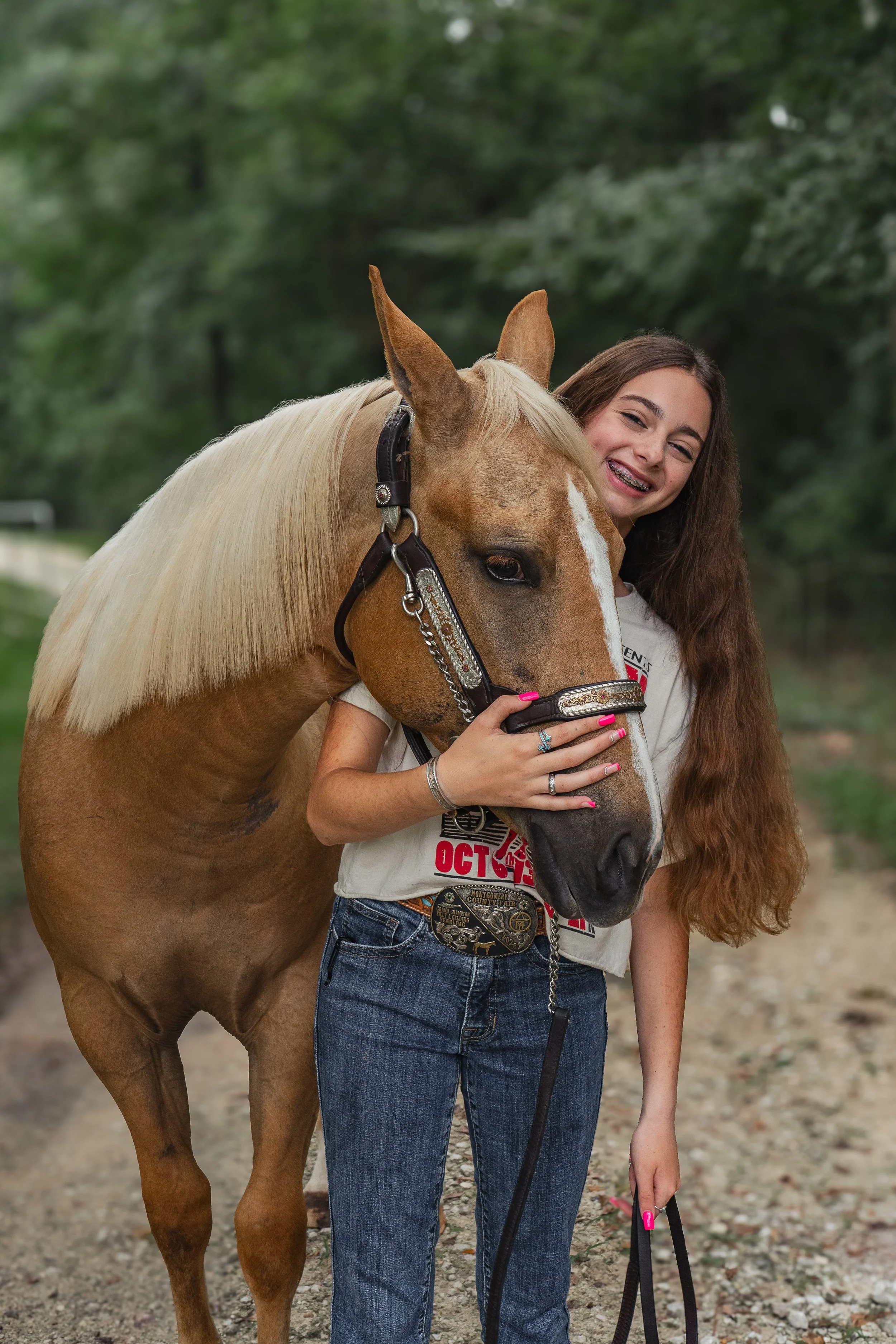 A girl with long curly hair and a big smile, wearing a white t-shirt and jeans, standing close to a brown horse with a light-colored mane outdoors. She is holding the horse's bridle with her right hand, and a pink leash with her left hand. The backgr