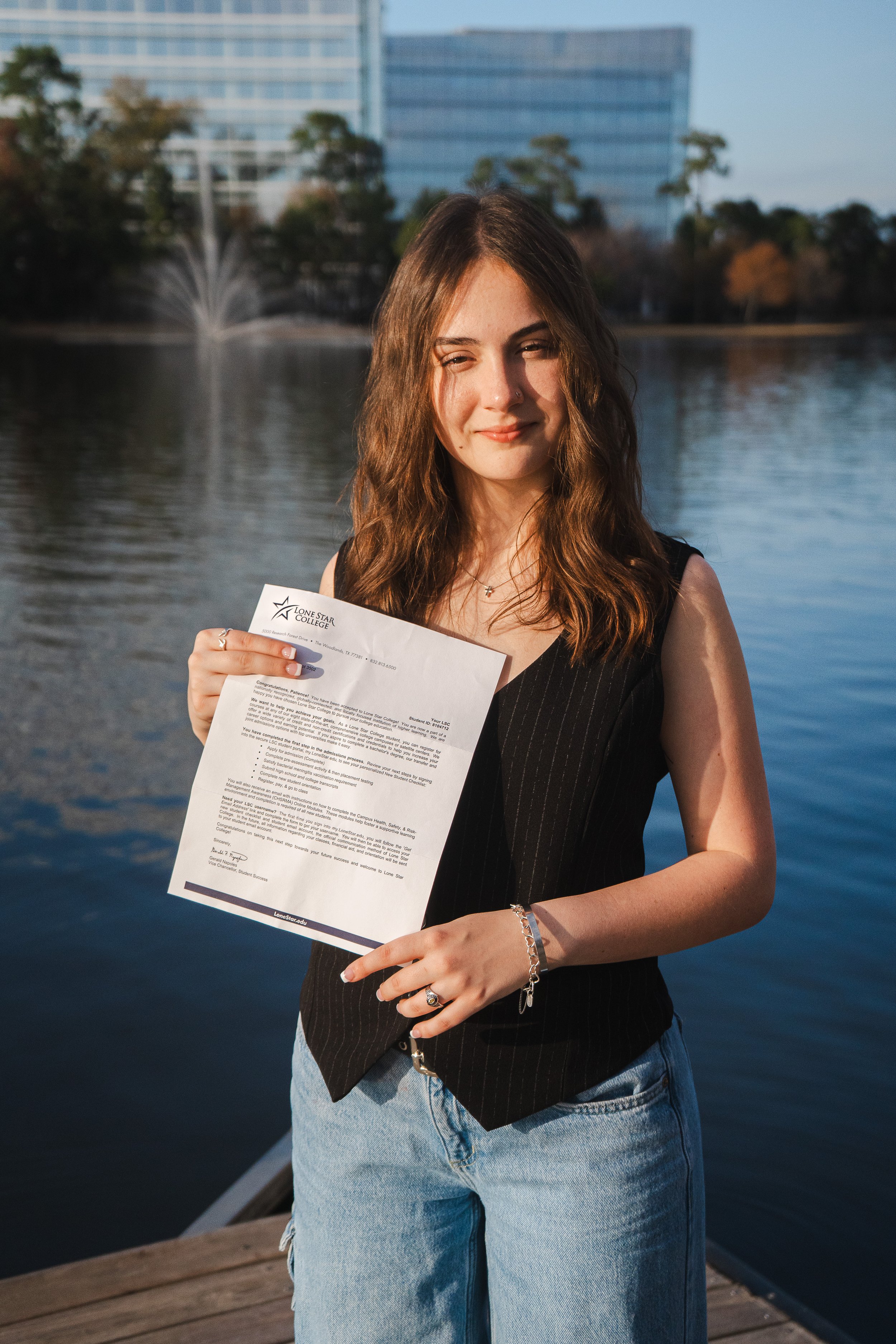Young woman holding a college acceptance letter in front of a water fountain and high-rise buildings at sunset.
