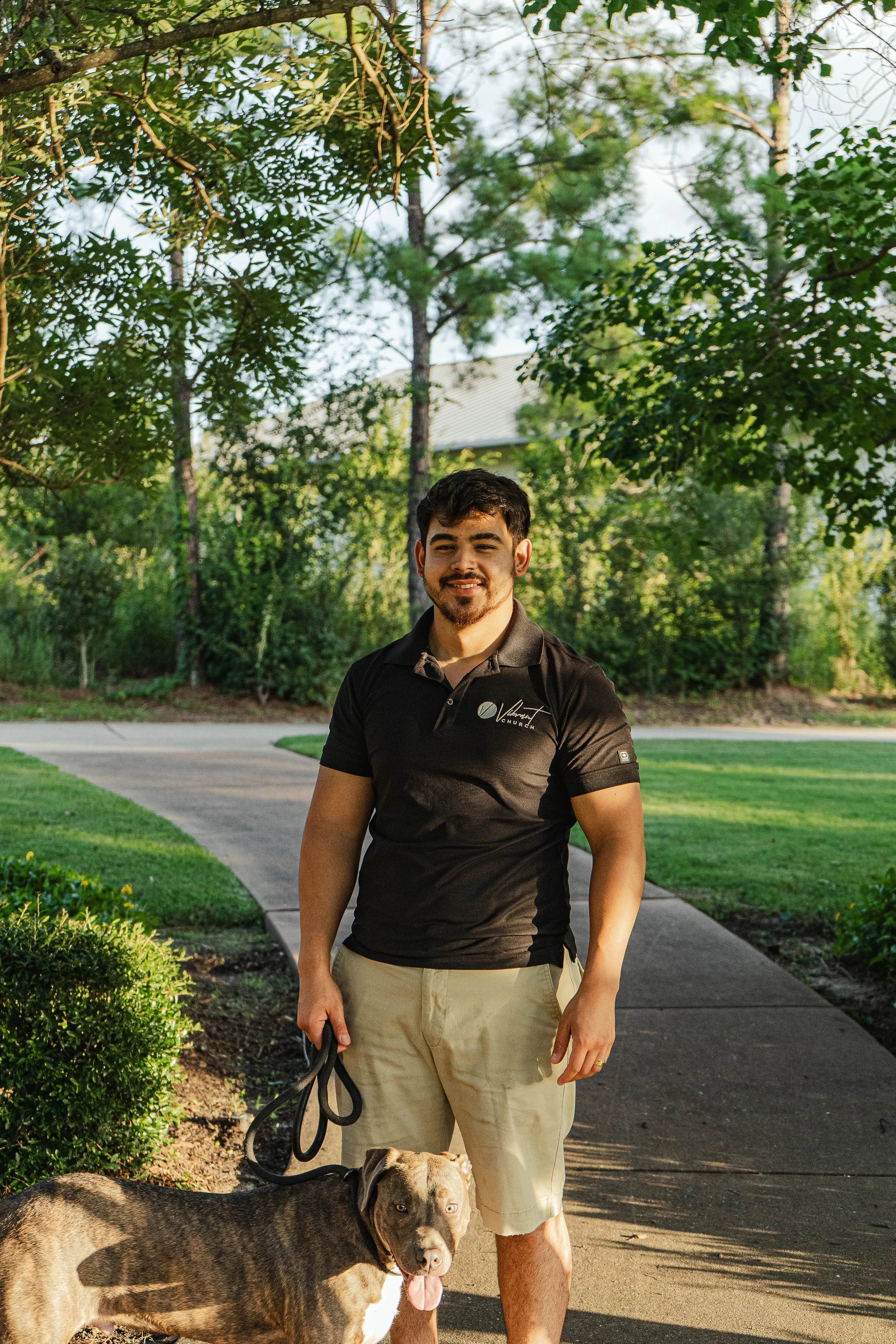 A young man with dark hair and a beard smiling outdoors on a sunny day, holding a leash attached to a large, gray and tan dog with a muscular build, standing on a sidewalk with green trees and bushes in the background.