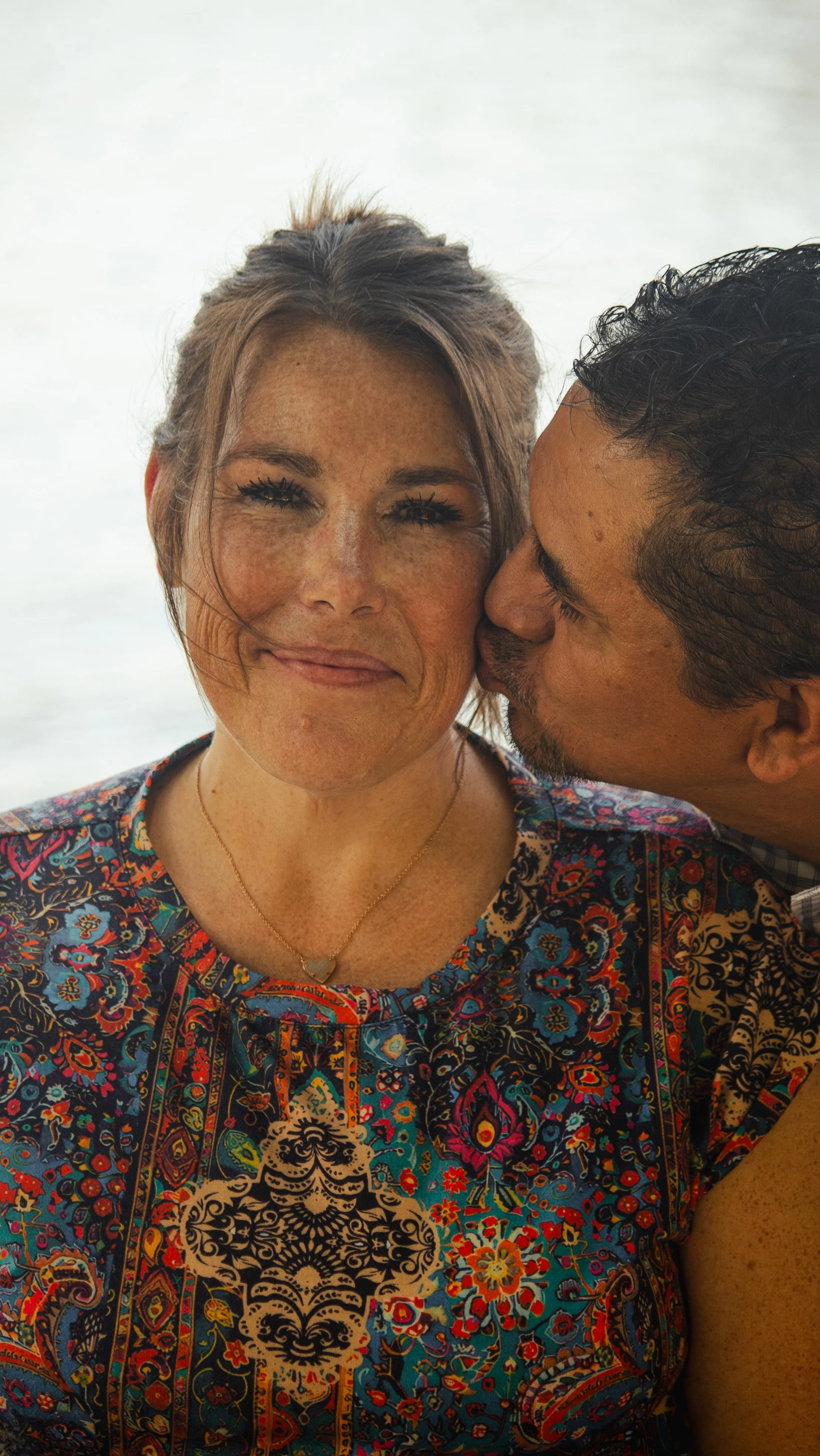 A woman with freckles smiling at the camera while a man kisses her cheek, both wearing colorful patterned clothing, in an outdoor setting.