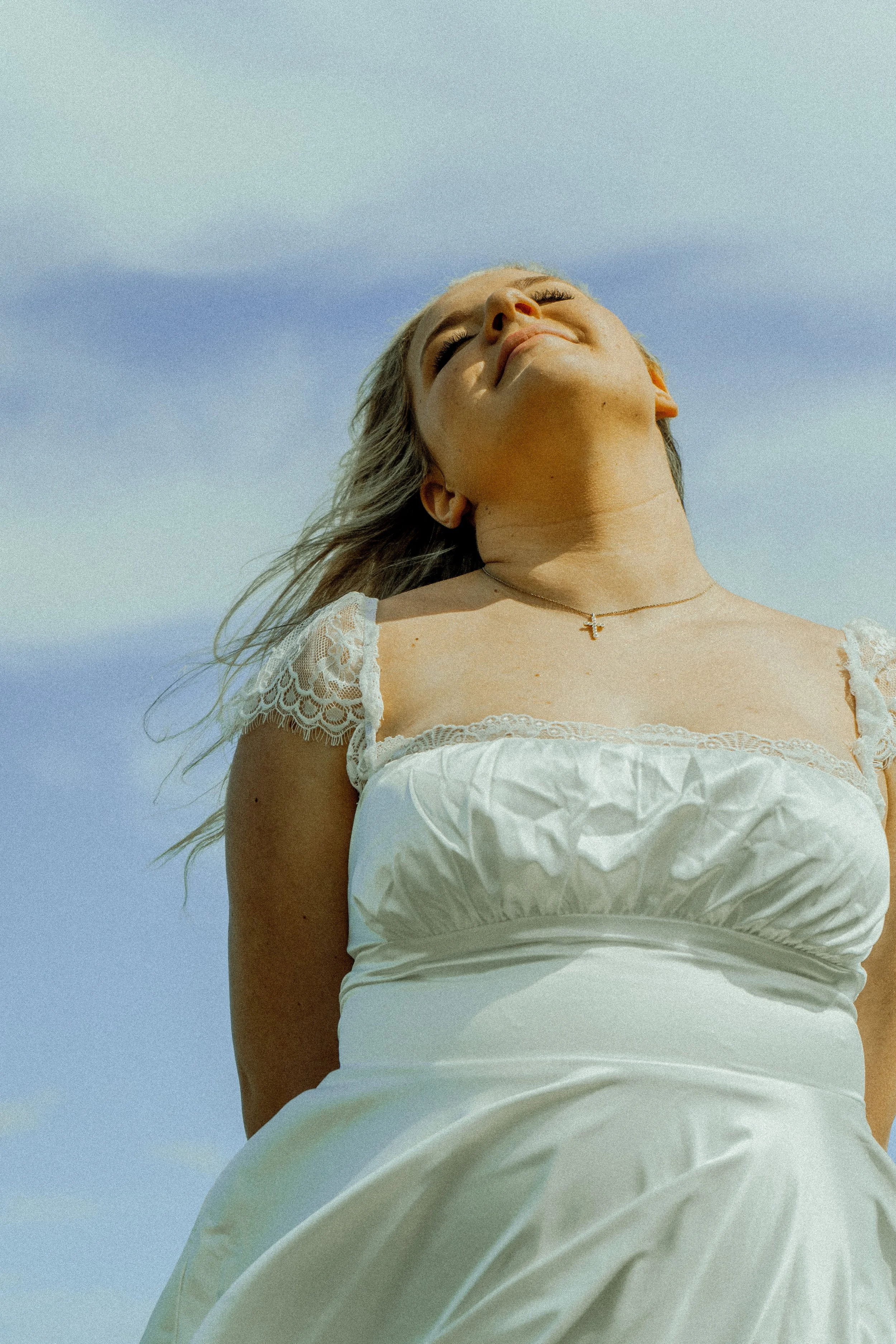 A woman with blonde hair and closed eyes smiling, wearing a white top with lace detail and a cross necklace, outdoors against a blue sky.
