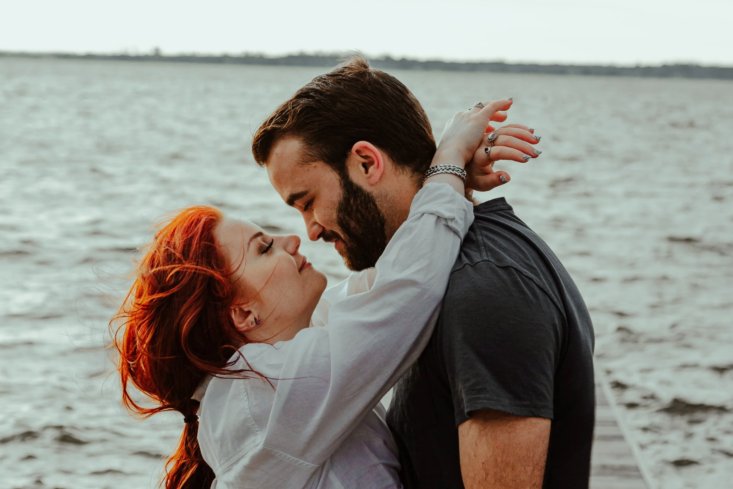 A man and woman with red hair embracing near a body of water, with the man’s face close to the woman’s face as they smile and look into each other's eyes.