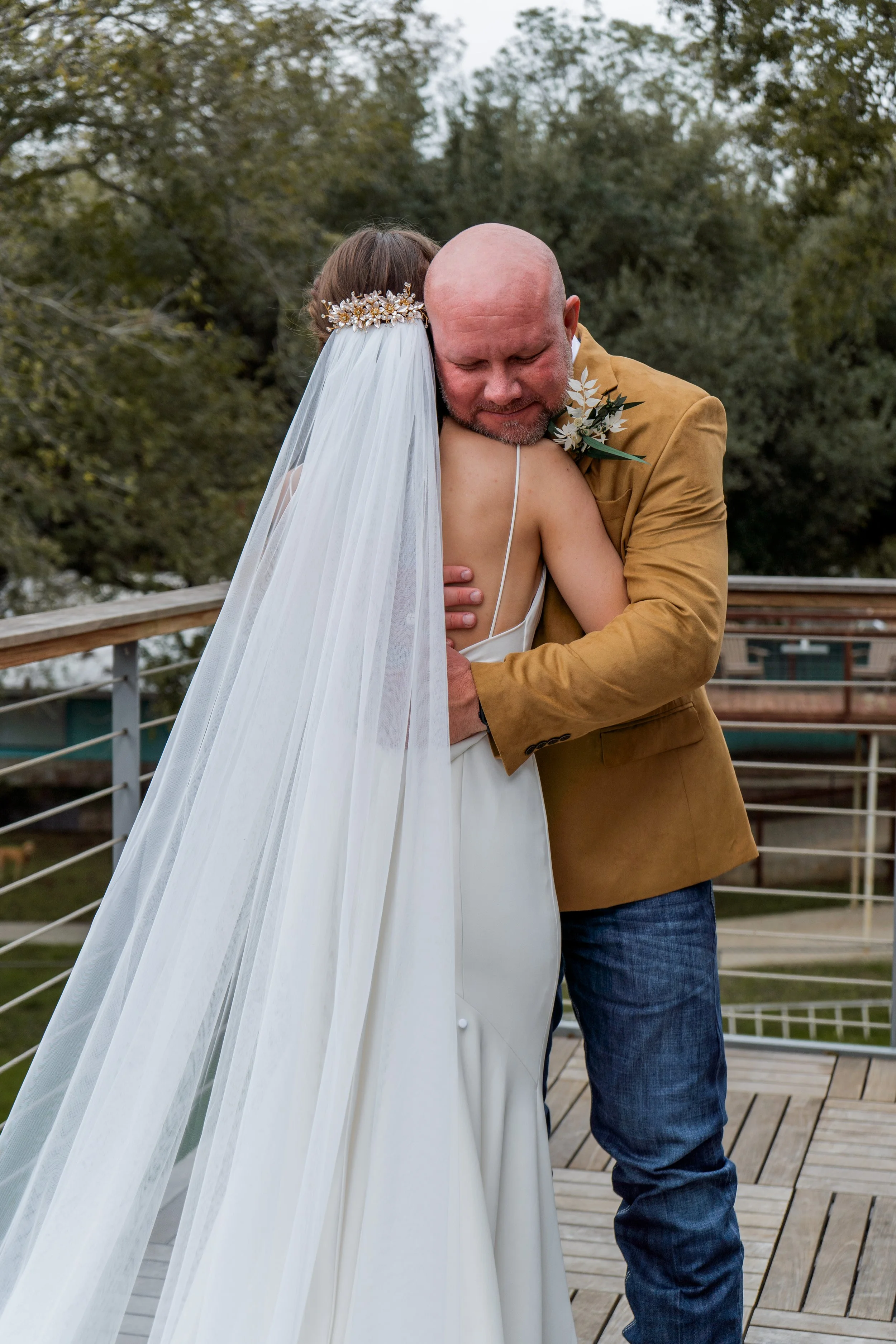 A bride and her father share a loving embrace during their first look outdoors on a wooden deck, with trees in the background.