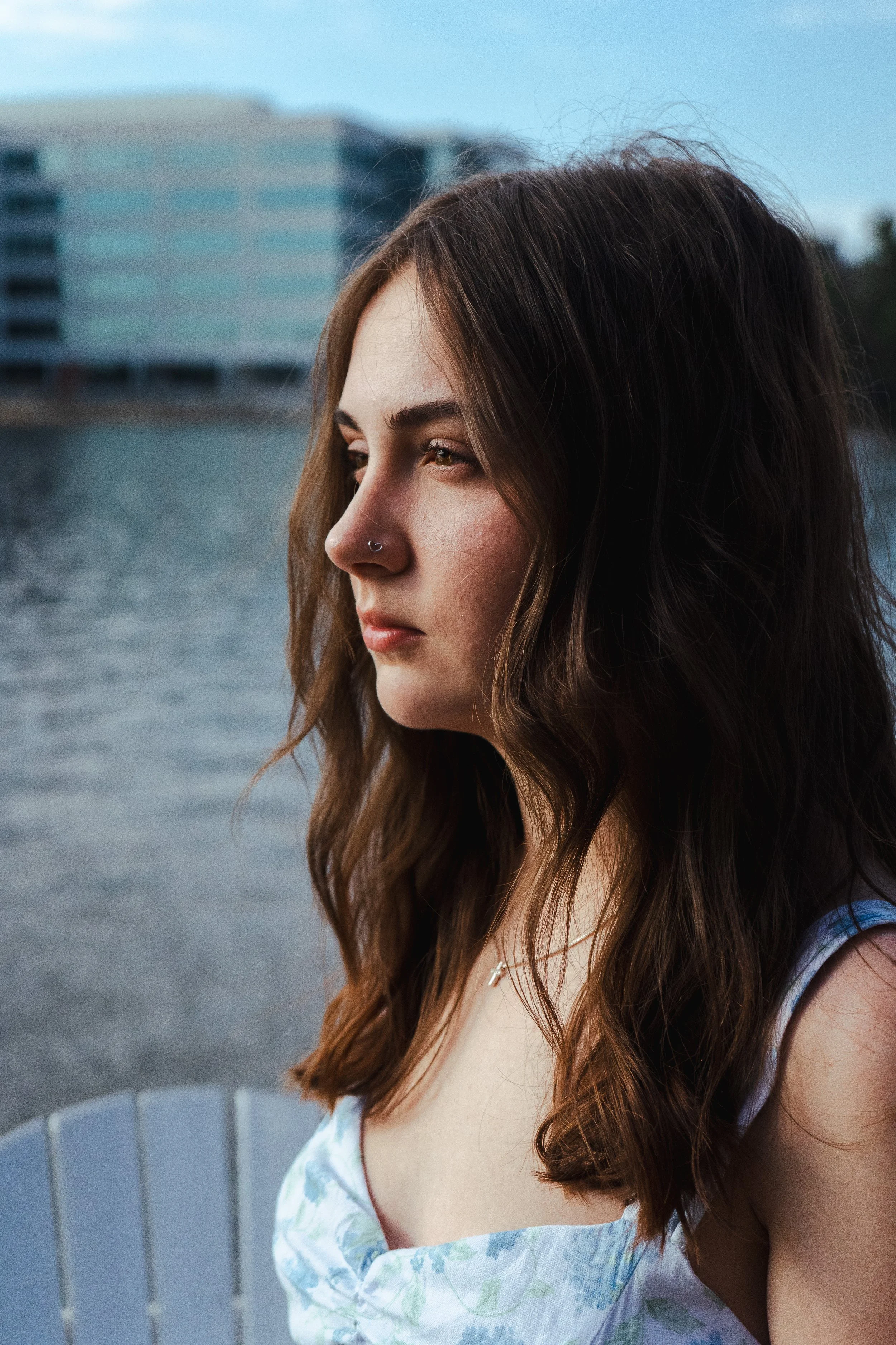 Close-up of a young woman with long brown hair and a nose piercing looking to the left, standing near water with a modern building in the background.